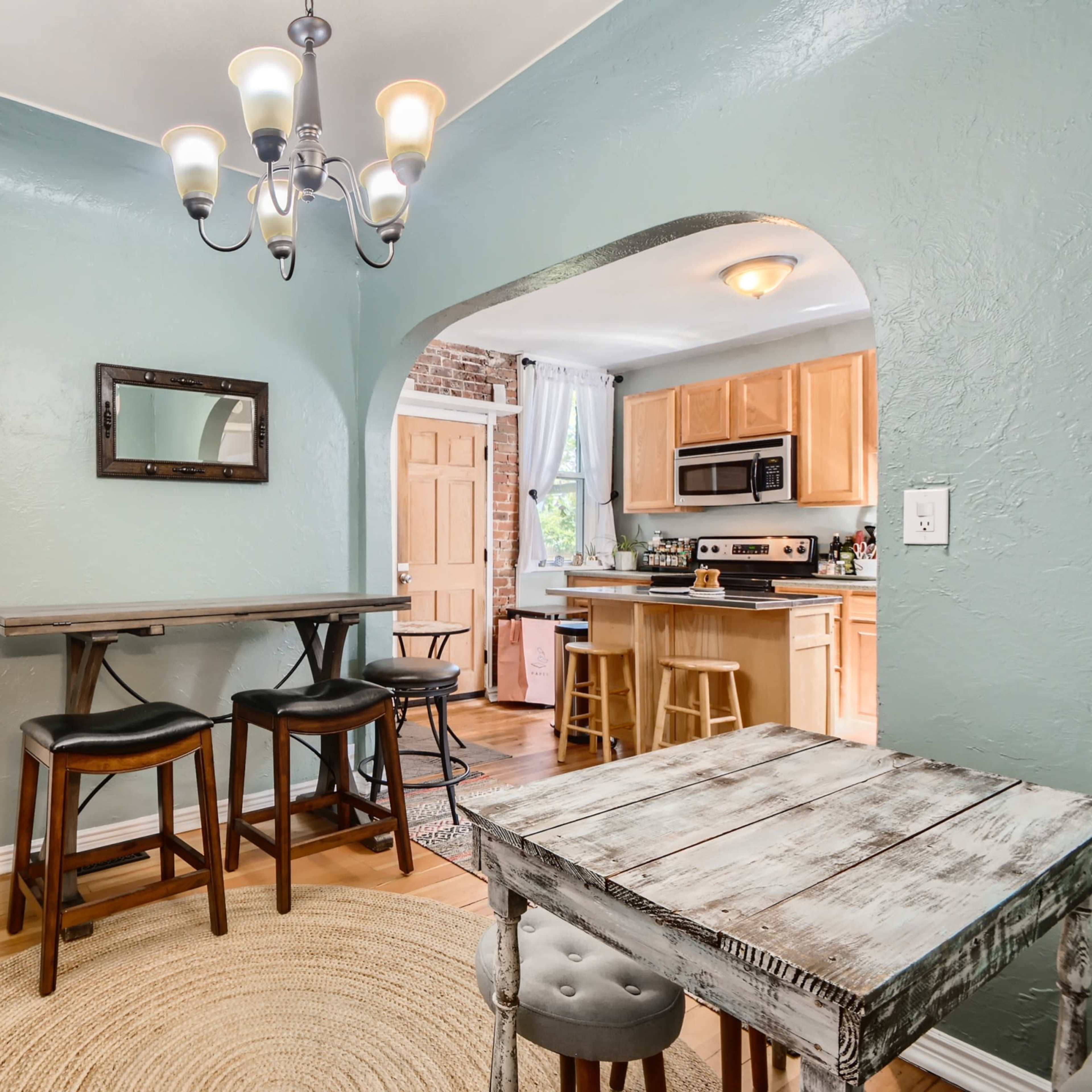 A dining area with a round rug, a wooden table with cushioned stools, and a small bar with high chairs, leading to a kitchen in the background.