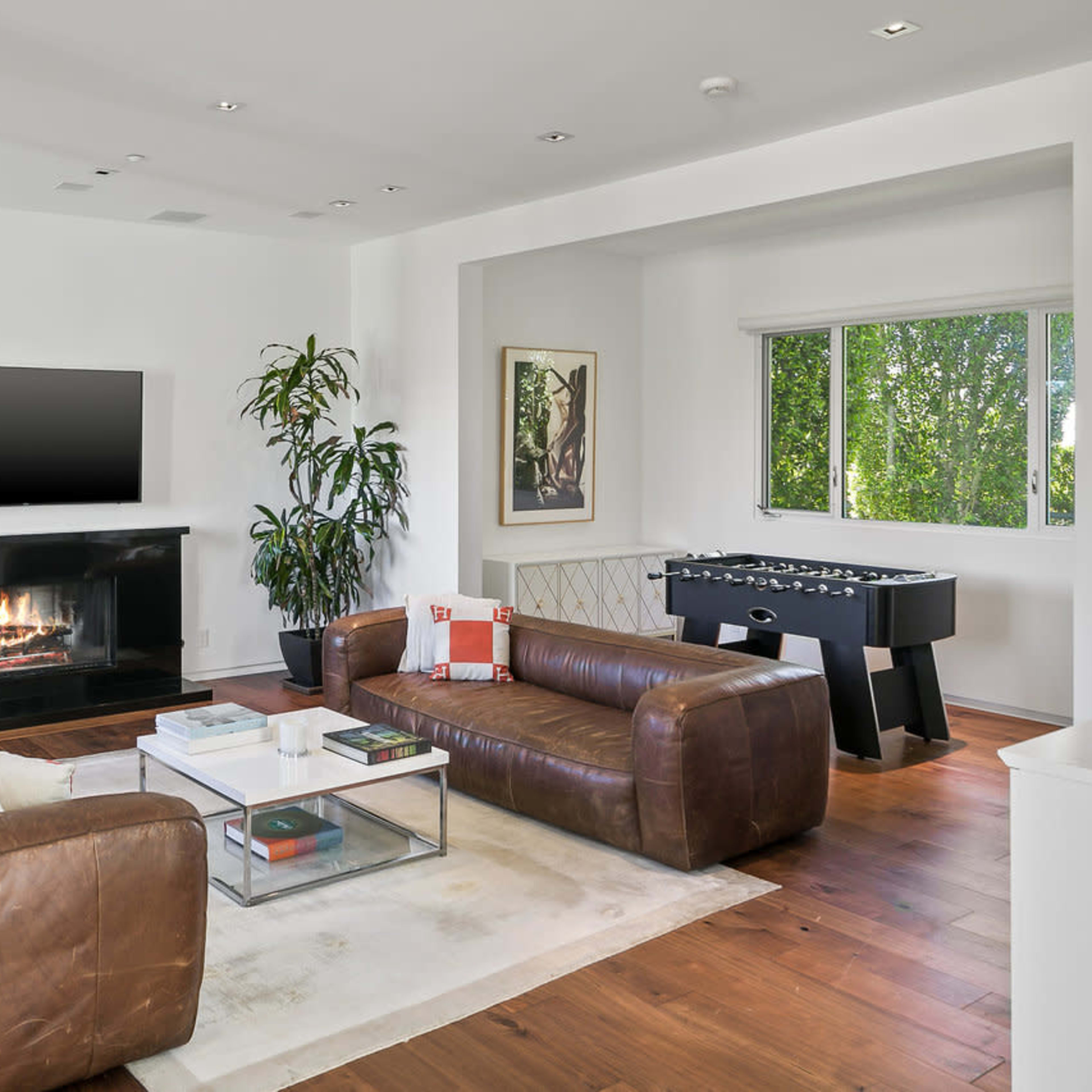 A bright living room features a leather sofa, a coffee table, a modern fireplace, and a foosball table near a window.
