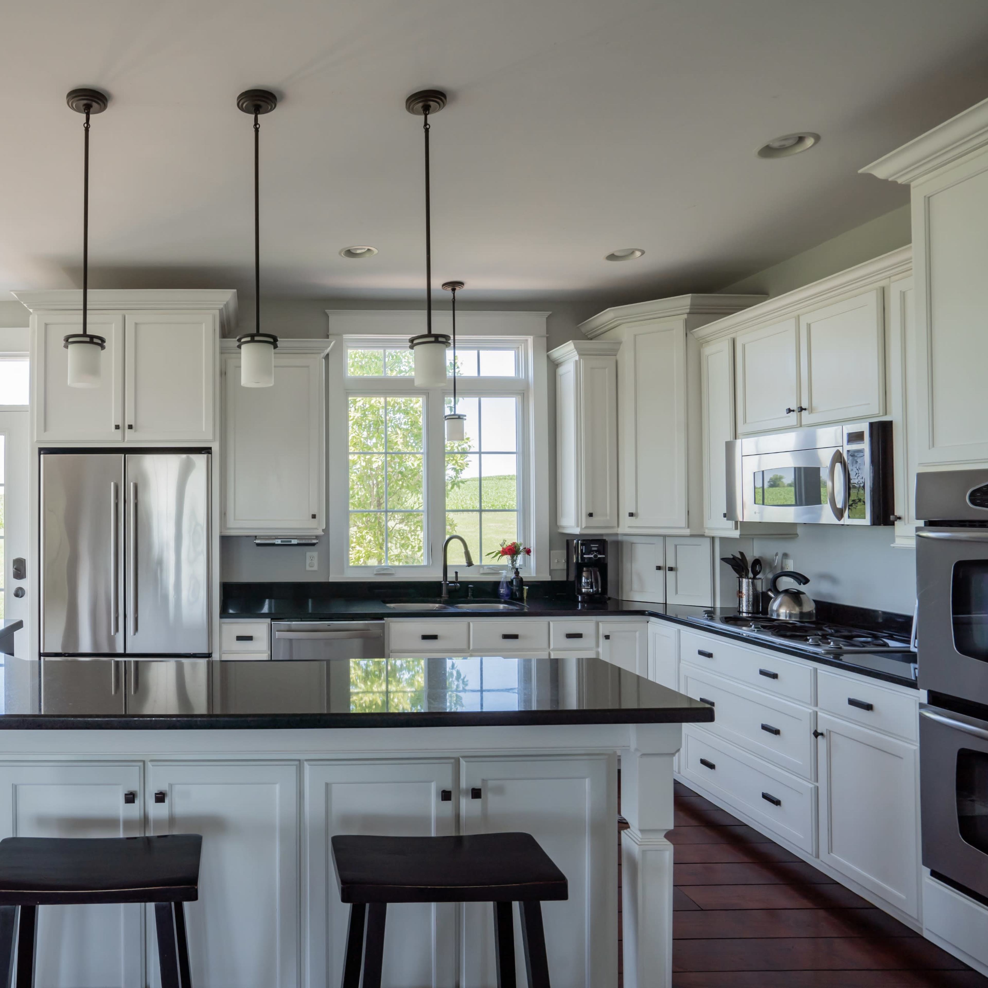A bright kitchen features white cabinets, a dark countertop, and large windows overlooking a garden.