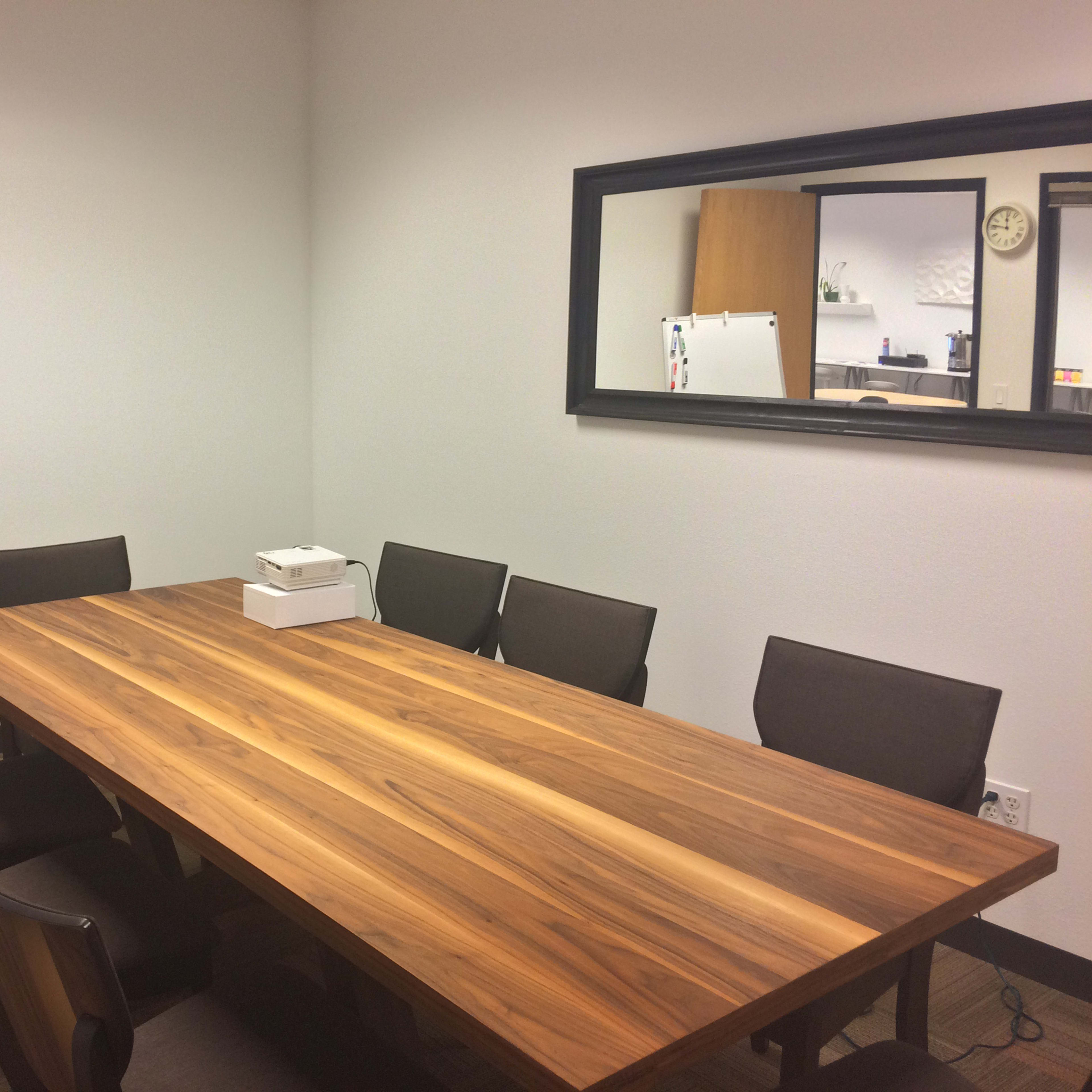 A conference room features a long wooden table surrounded by eight chairs, with a whiteboard and a mirror on the walls.