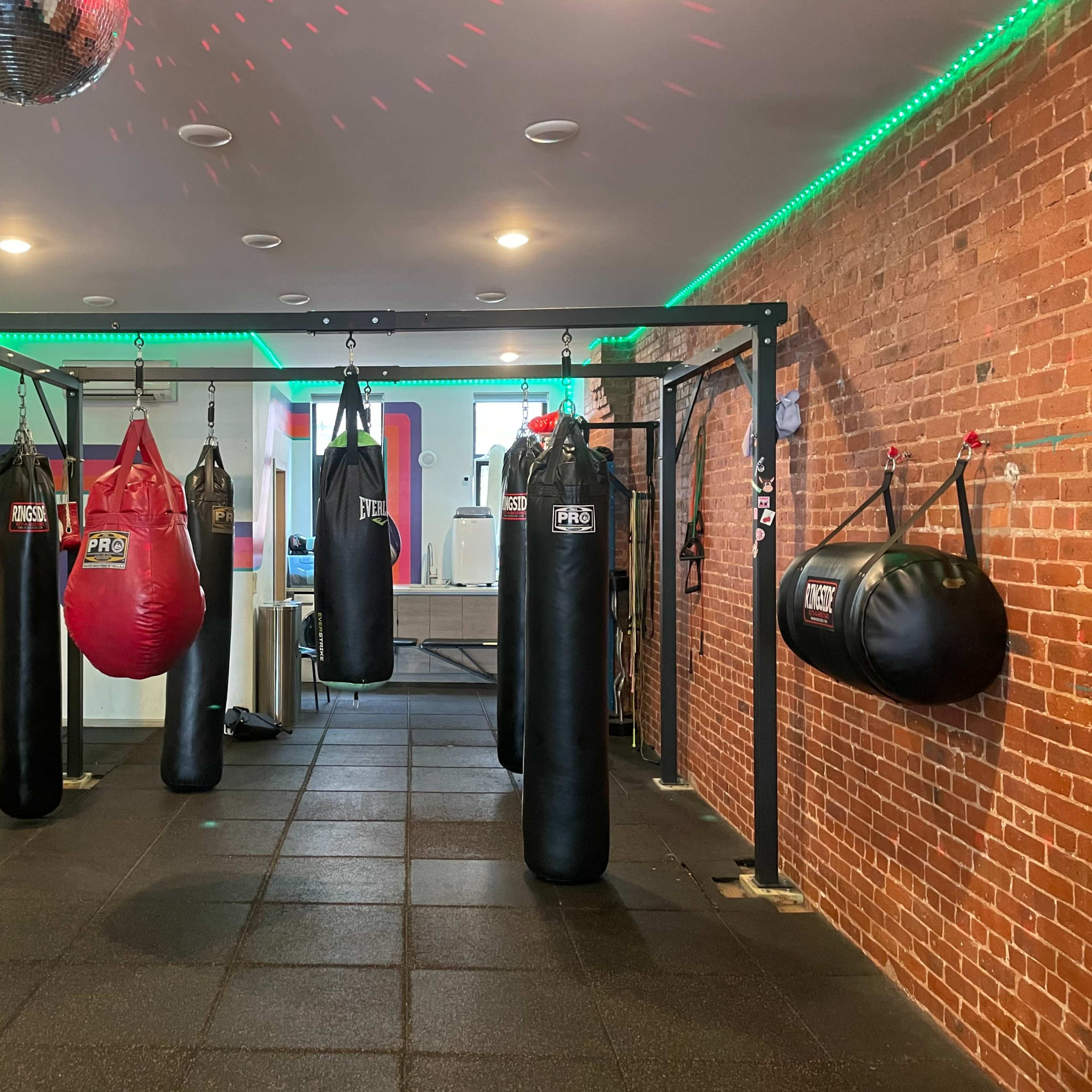 A boxing training area with multiple punching bags hanging from a metal frame, surrounded by a brick wall and colorful wall decorations.