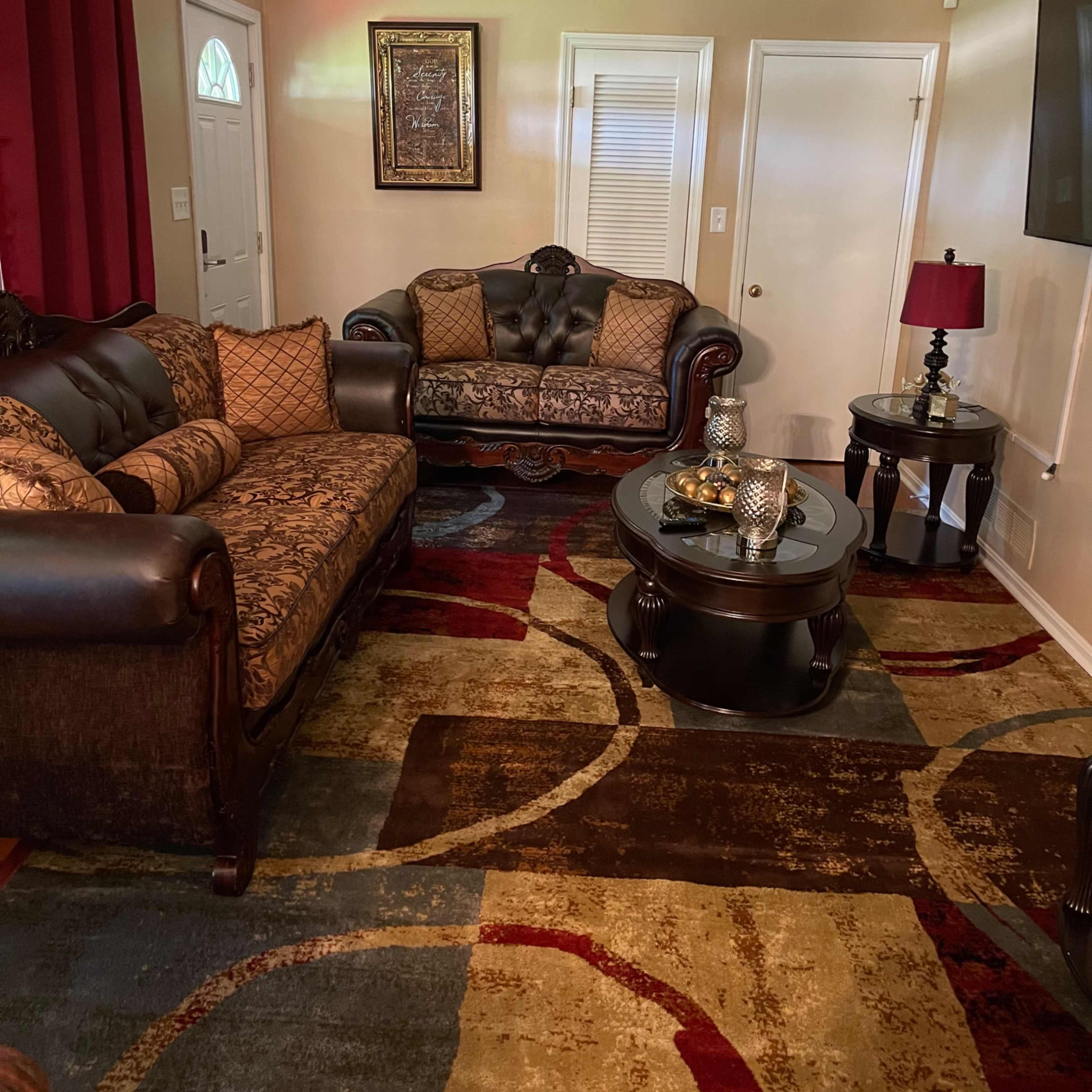A living room with a brown leather sofa and loveseat, a round glass coffee table, and a colorful rug with circular patterns.