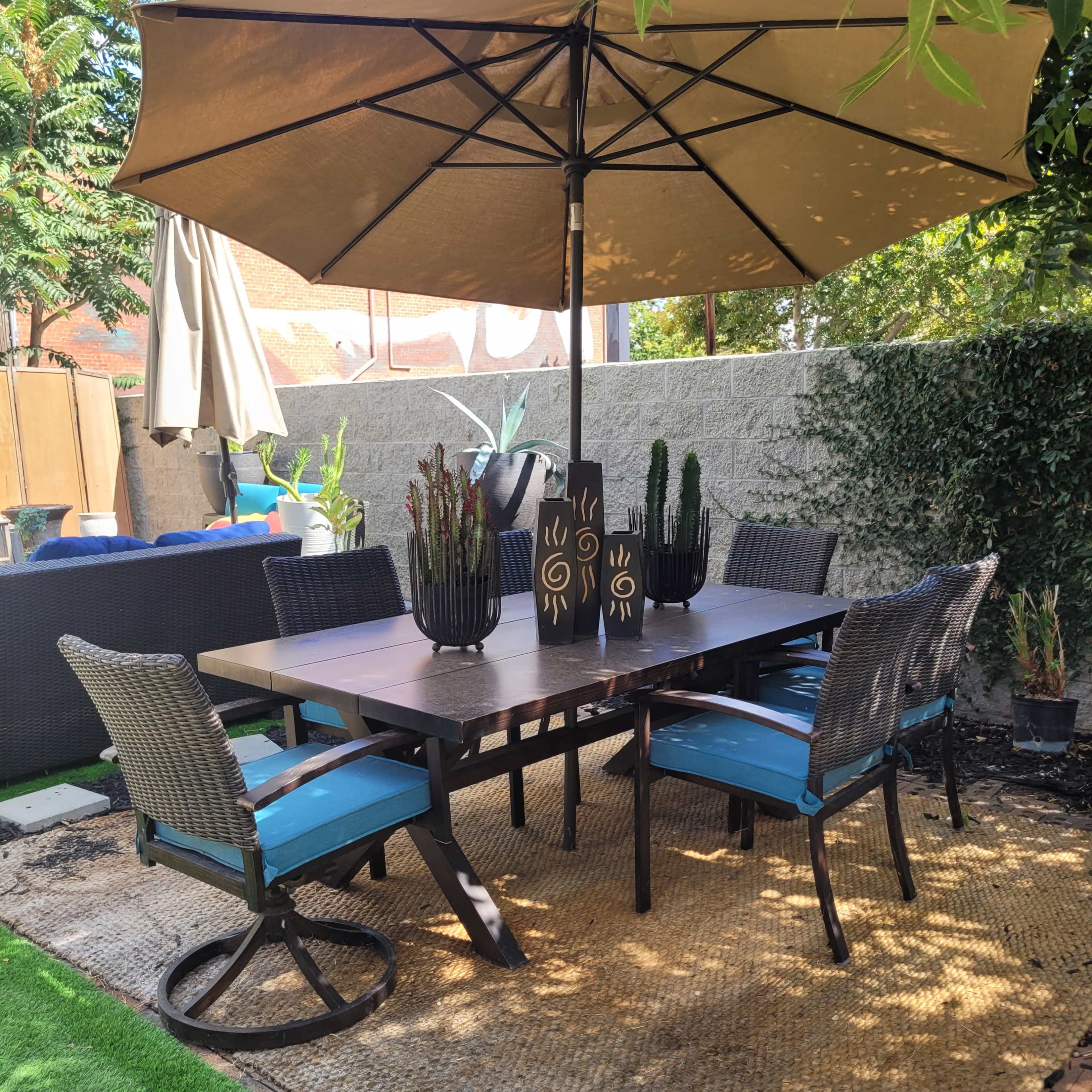 A patio area features a dining table with six chairs under a large umbrella, surrounded by plants and landscaping.