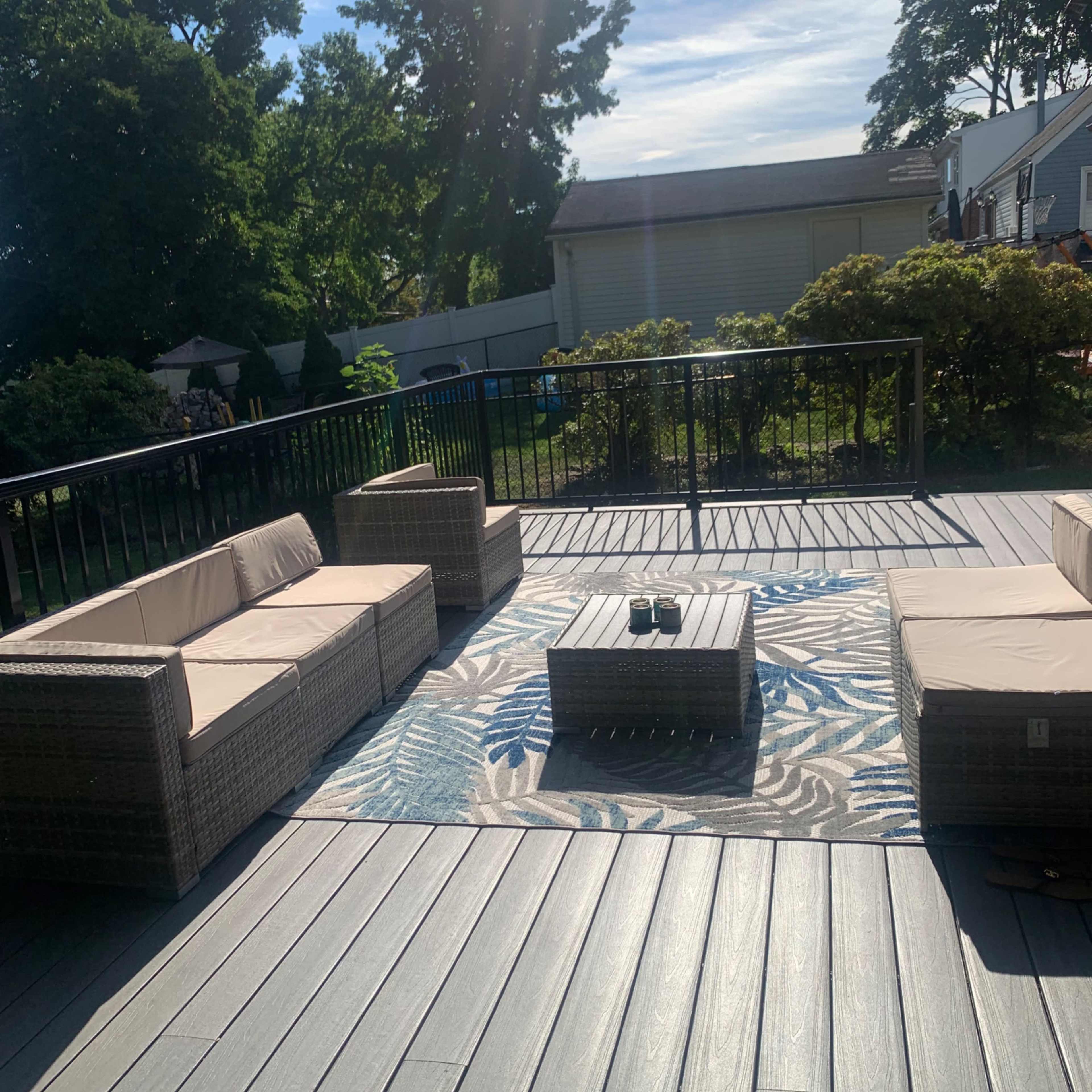 An outdoor seating area with a light-colored couch set and a coffee table on a deck, surrounded by greenery and a blue sky.