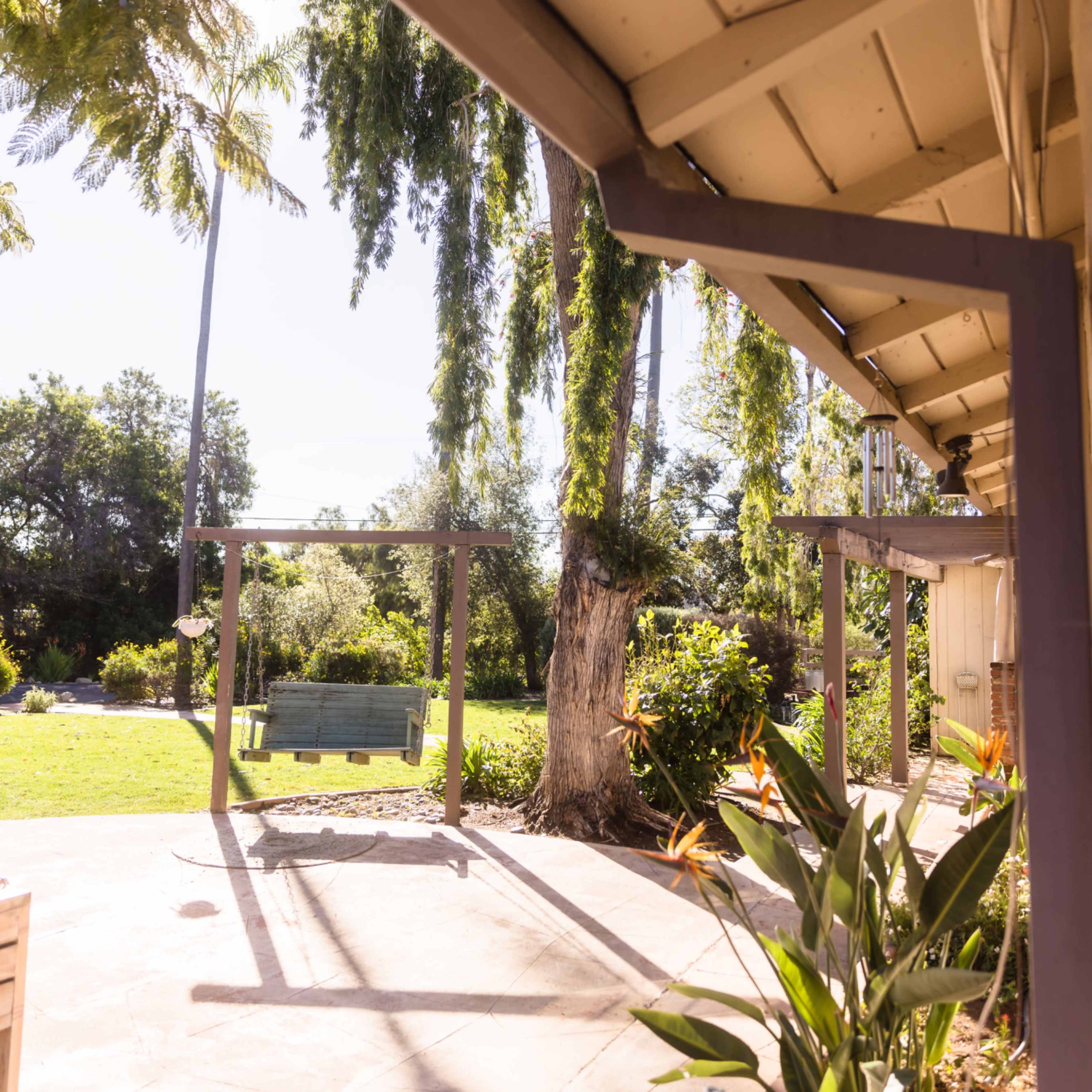 A patio area with a sloped roof, surrounding greenery, and a bench in a sunny garden.