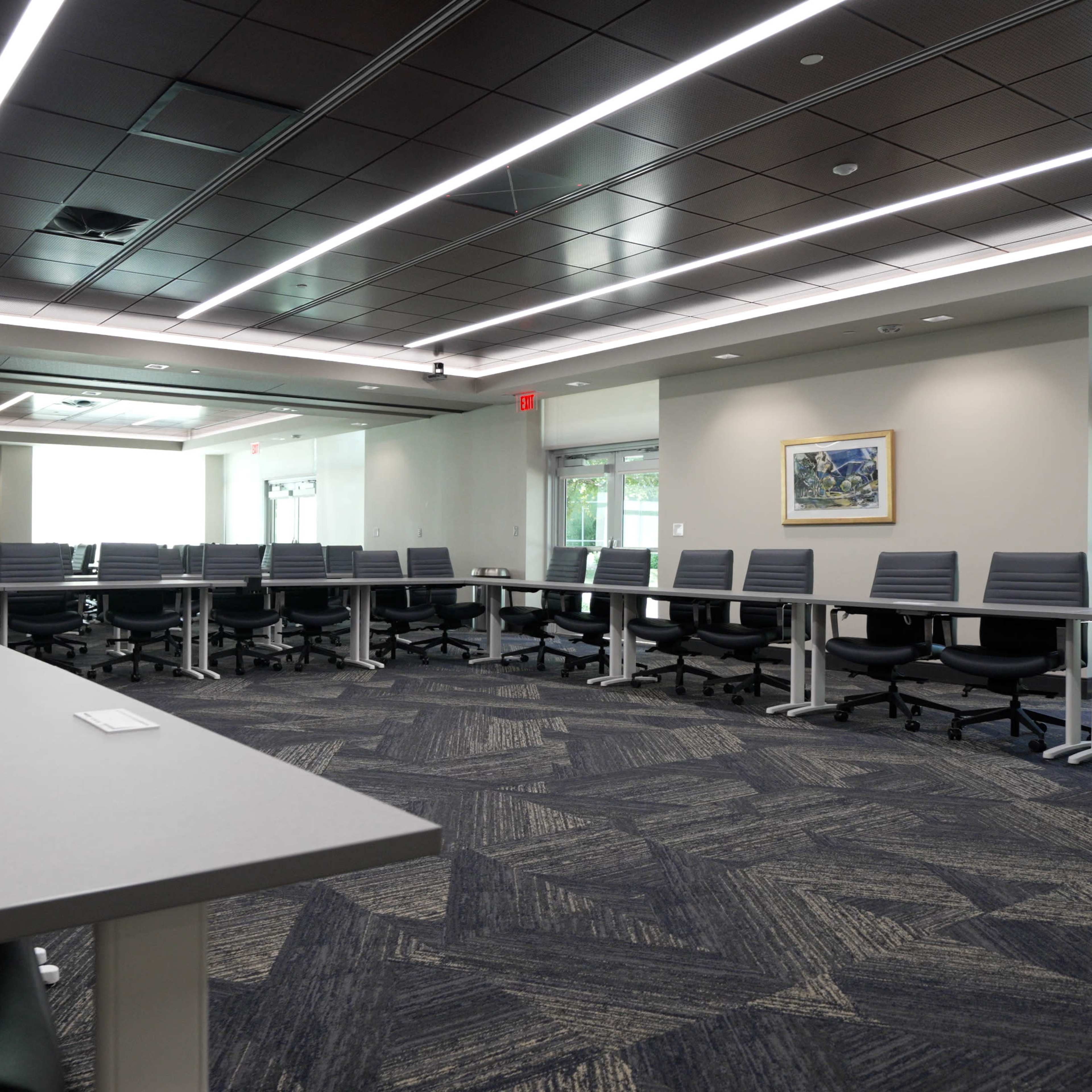 A modern conference room with a long rectangular table, multiple office chairs, and large windows providing natural light.