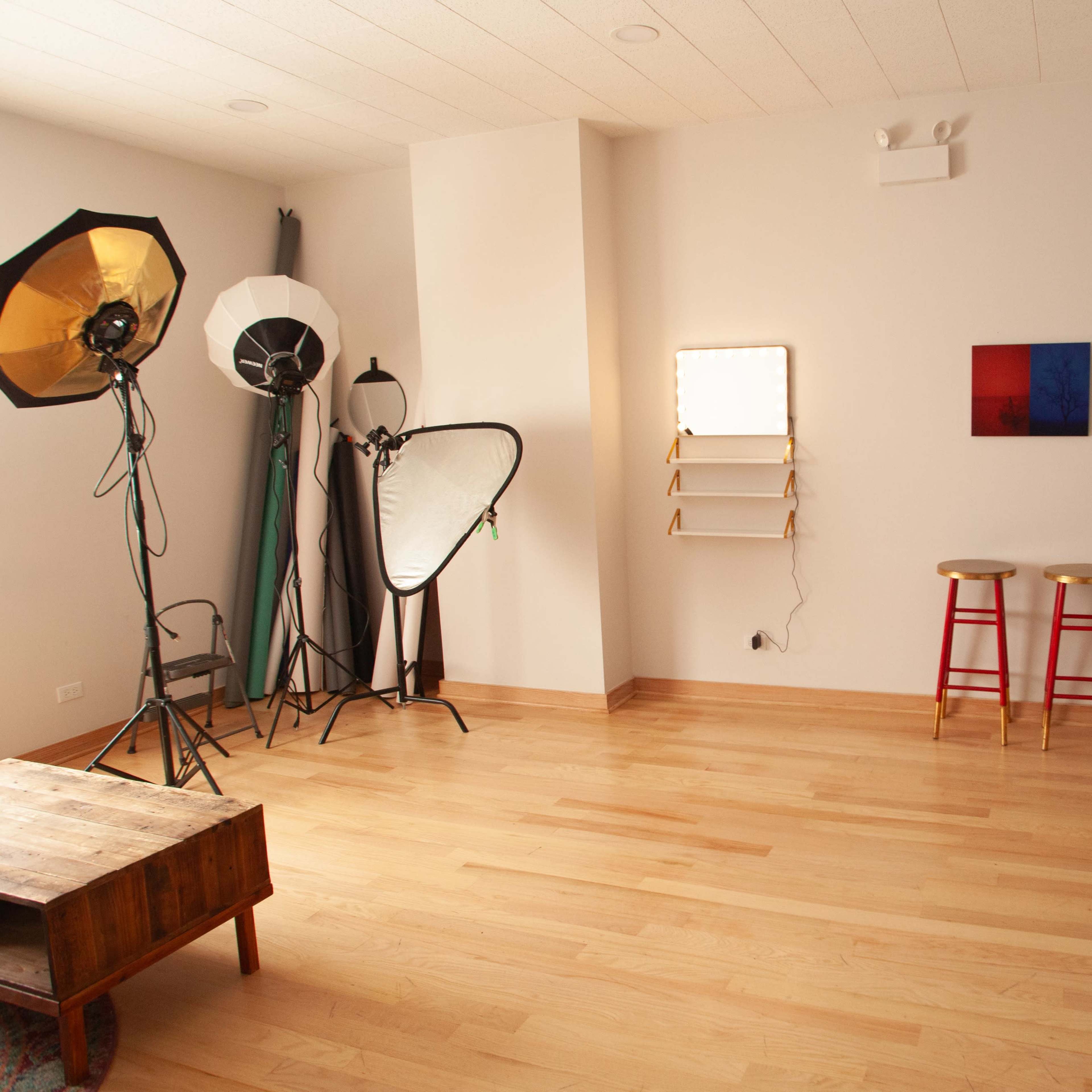 A photography studio with lighting equipment, a backdrop, two red stools, and a wooden coffee table on a hardwood floor.