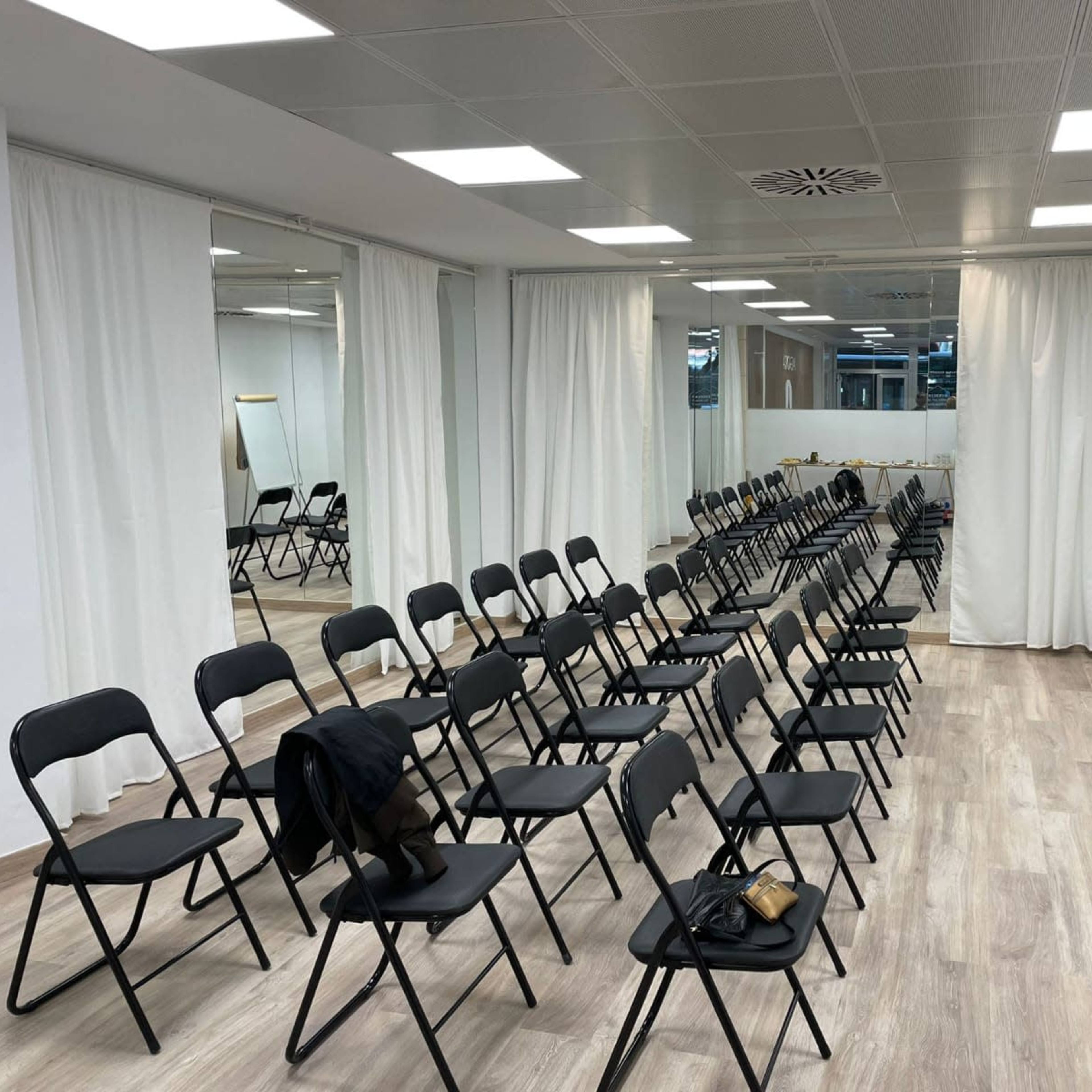 A conference room is set up with rows of black chairs facing a whiteboard, surrounded by white curtains and a light-colored floor.