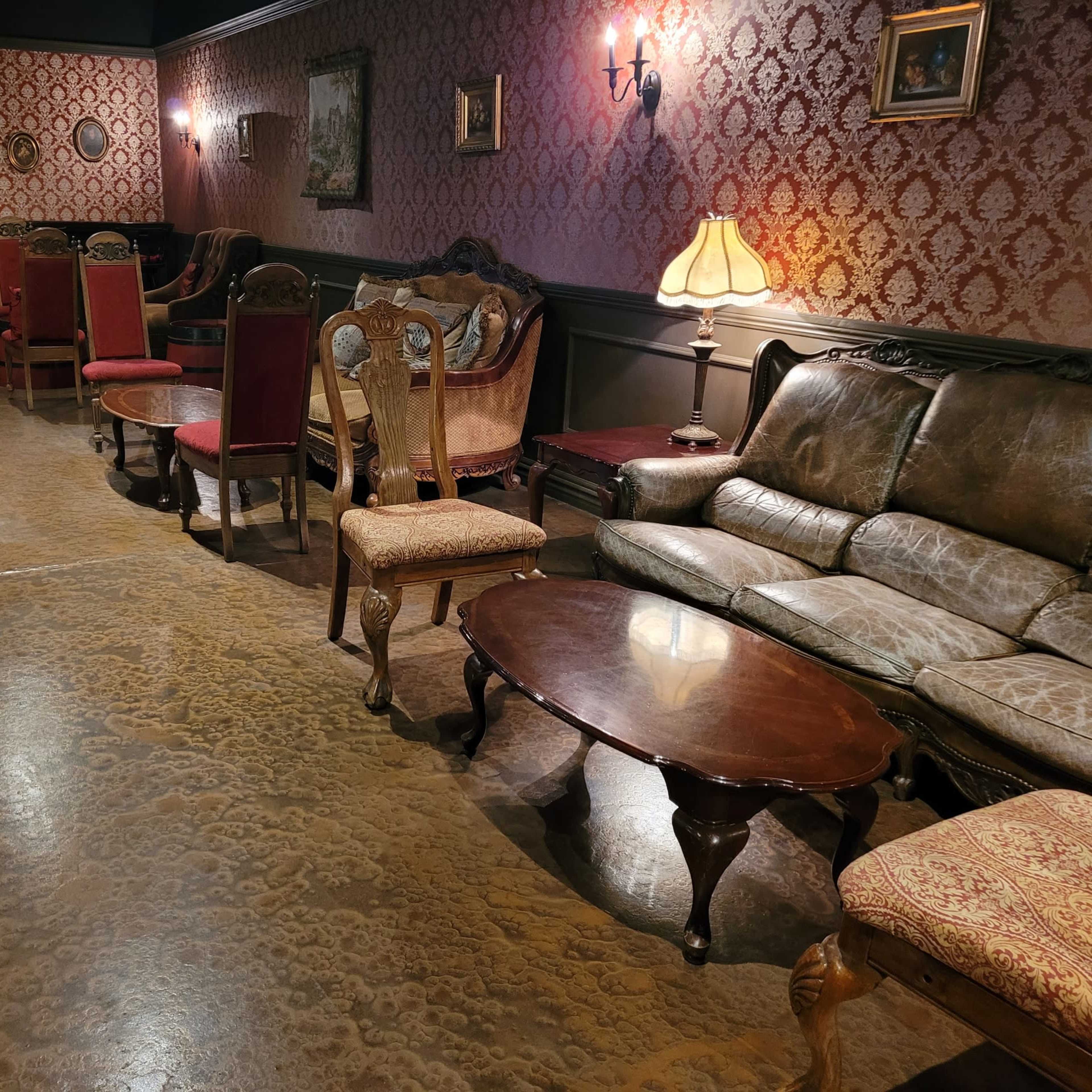 A cozy lounge area with vintage furniture, including leather sofas and wooden chairs, set against ornate wallpaper and dim lighting.