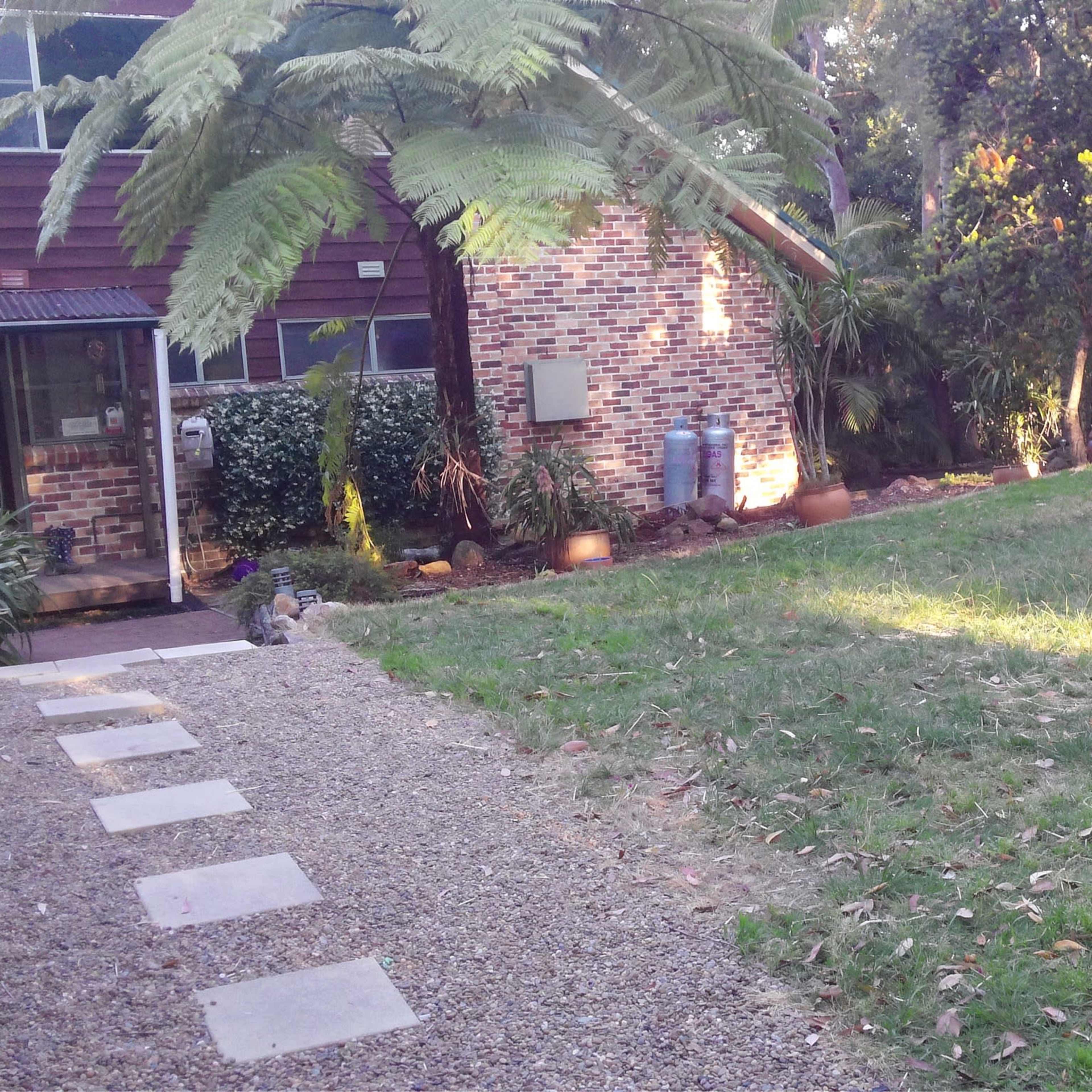 A brick house in a wooded area, with a stone path leading to a grassy lawn on one side and a small wooden structure in the background.