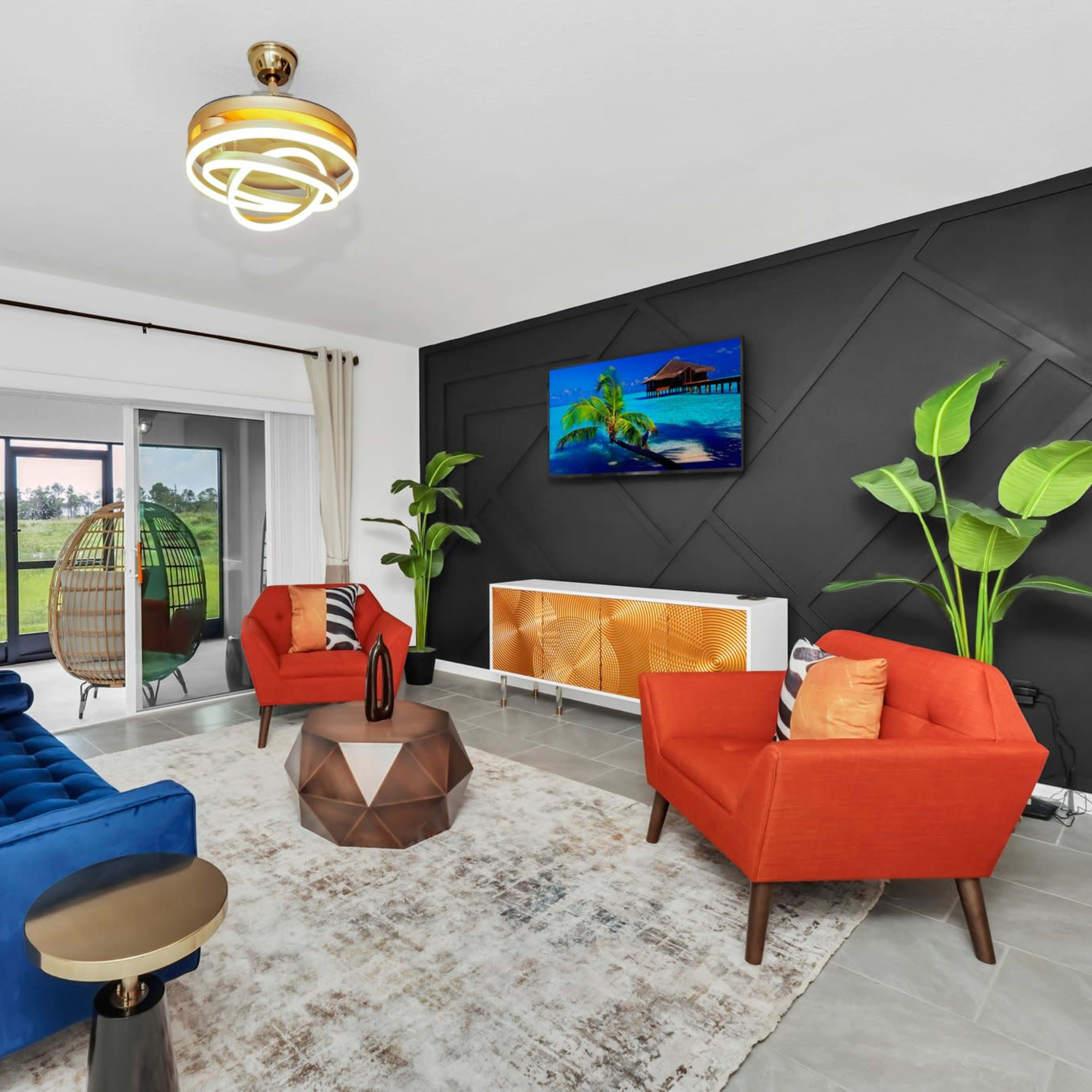The living room features two orange chairs and a blue sofa centered around a geometric coffee table, with a large TV mounted on a dark accent wall and a view of greenery through sliding glass doors.