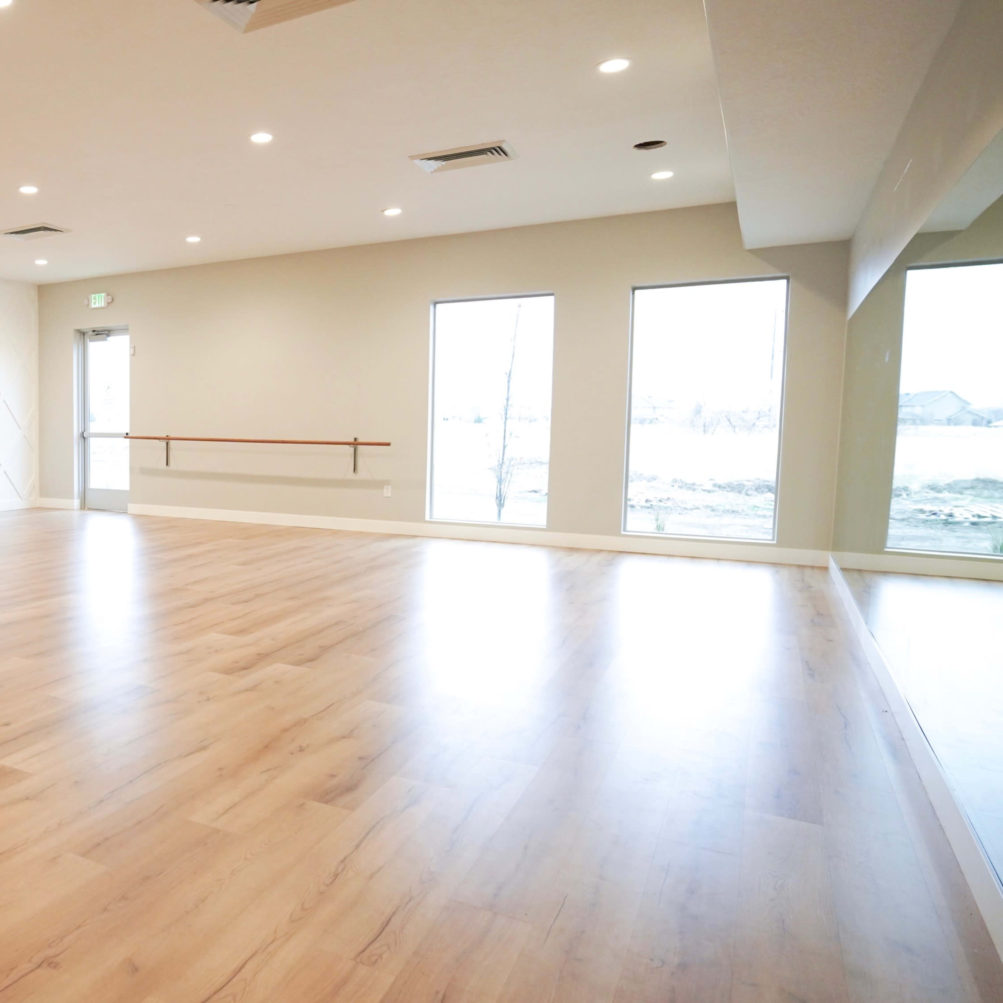 A spacious room with polished wooden flooring, a textured white wall, and large windows allowing natural light to enter.