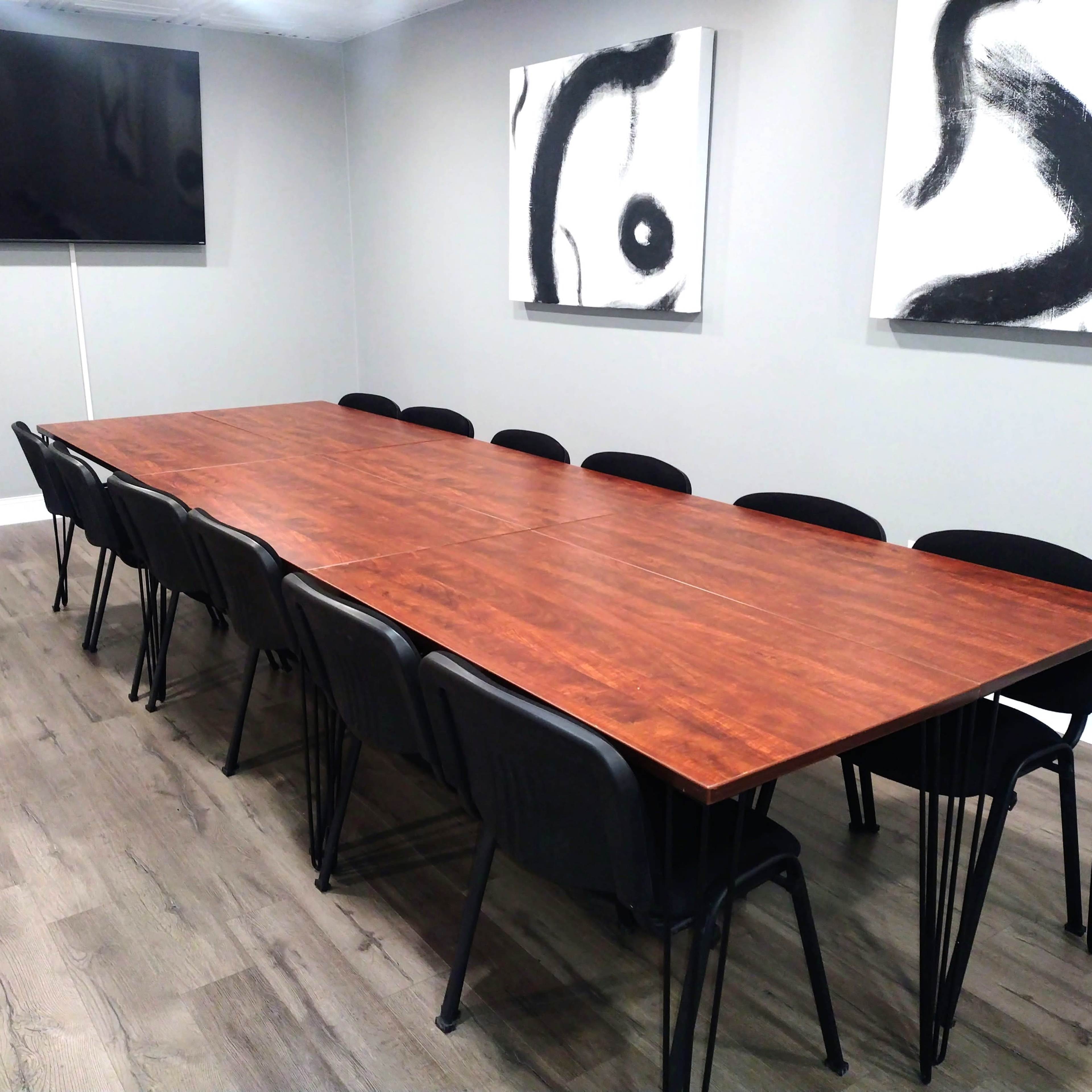 A long wooden table with black chairs is set in a meeting room featuring a large television and abstract black-and-white artwork on the walls.