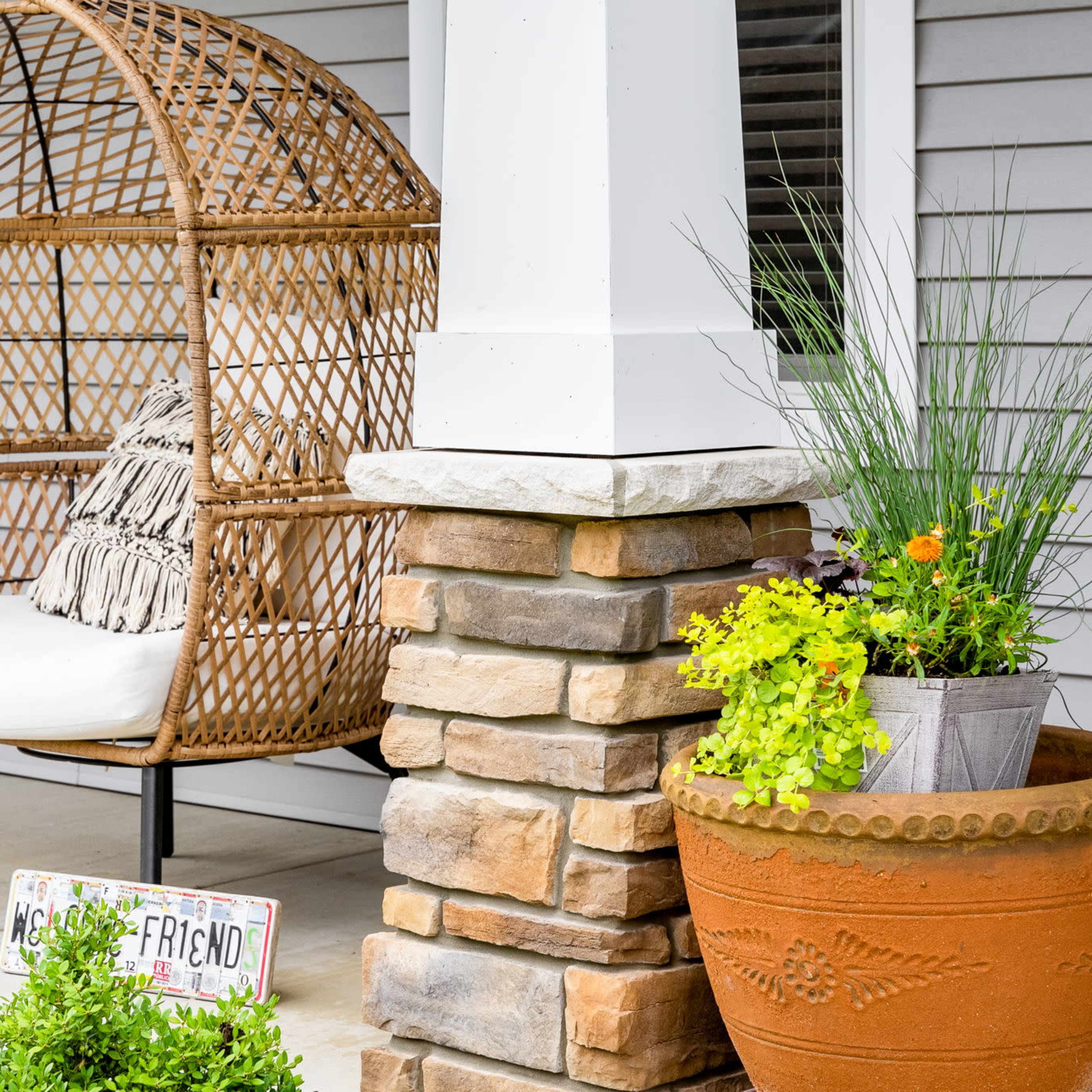 A woven basket chair with a cushion sits on a porch beside potted plants and stone pillars.