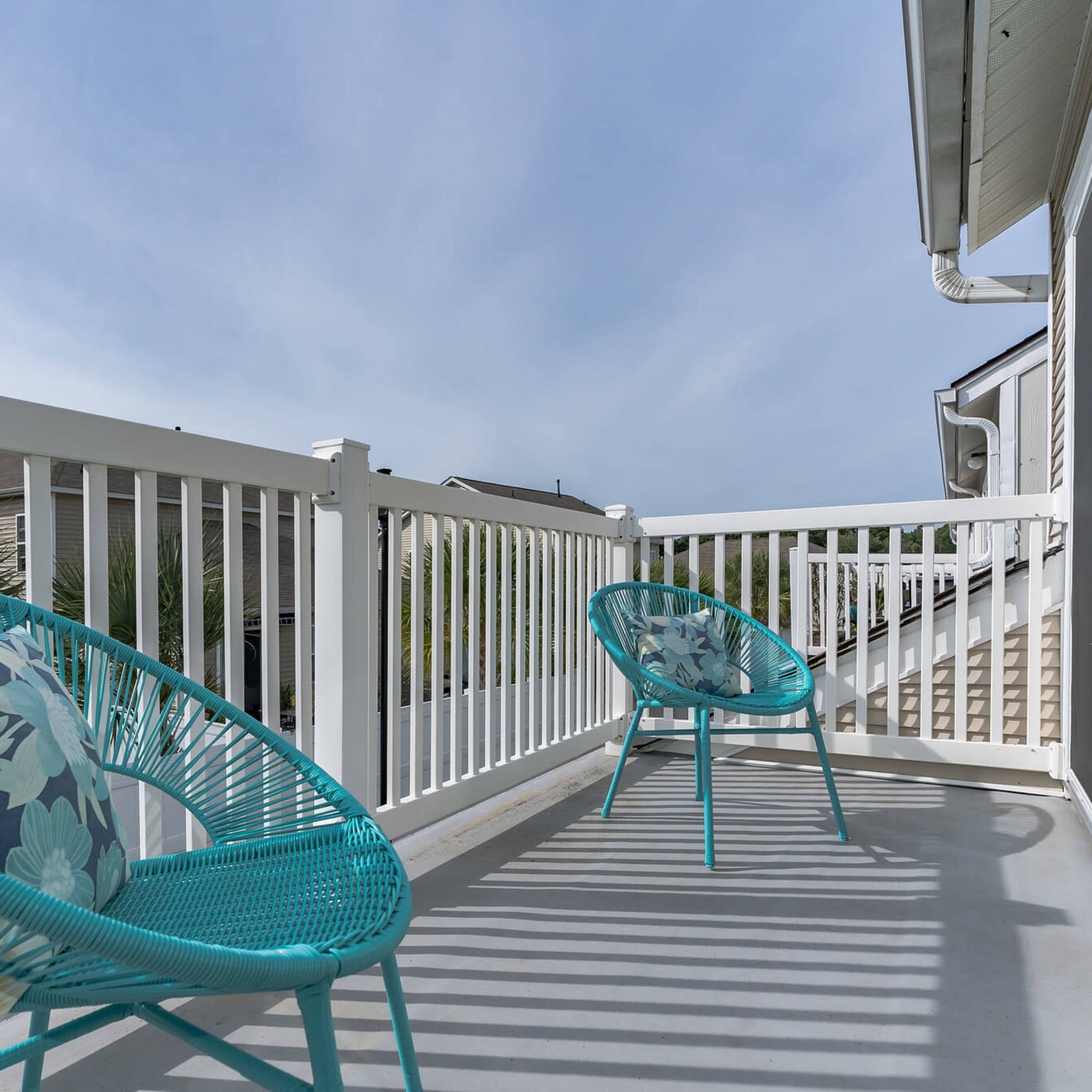 A balcony with two turquoise chairs and decorative pillows, featuring a view of nearby buildings and palm trees.