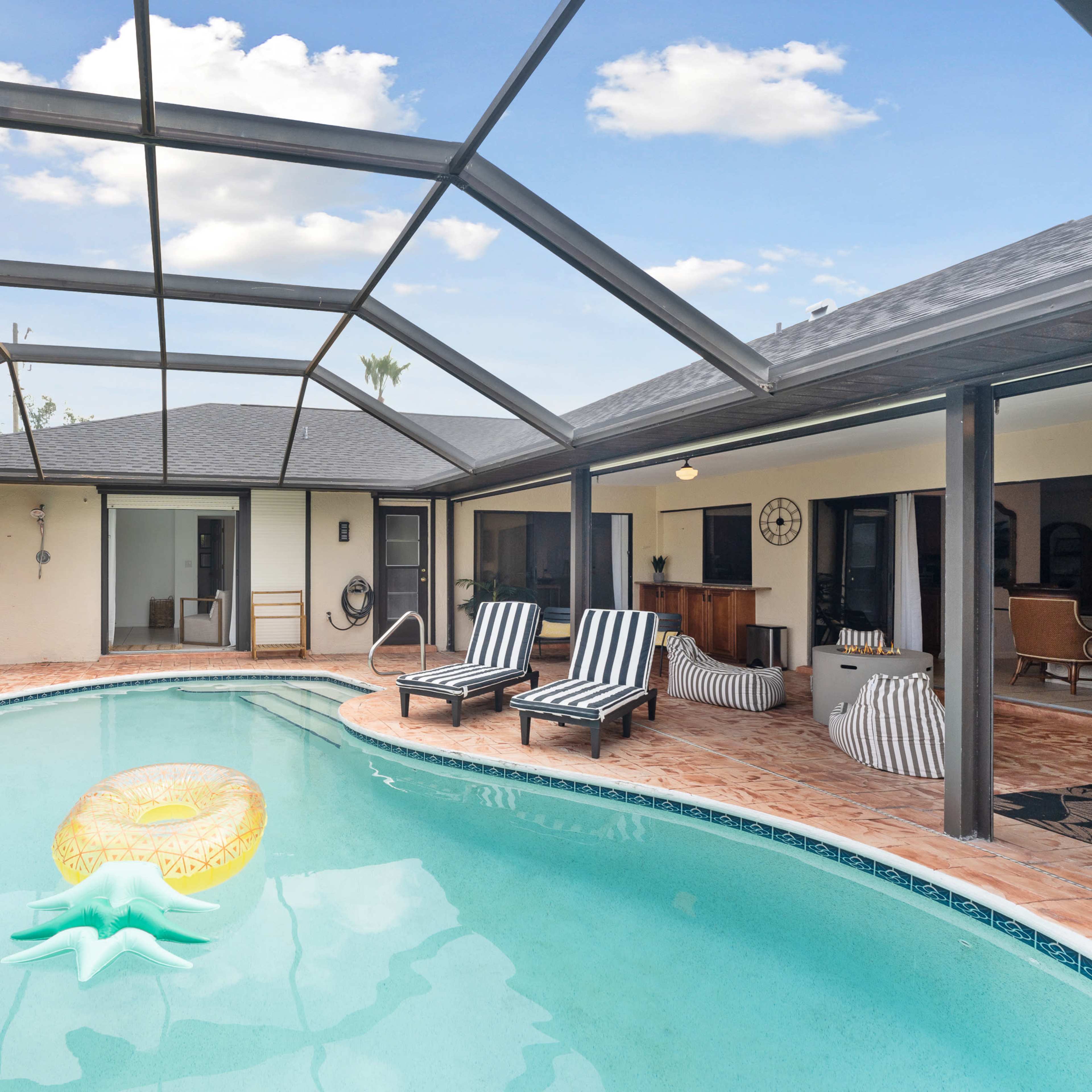 A screened-in pool area featuring a pineapple-shaped float, two lounge chairs, and a view of a modern home with large windows.