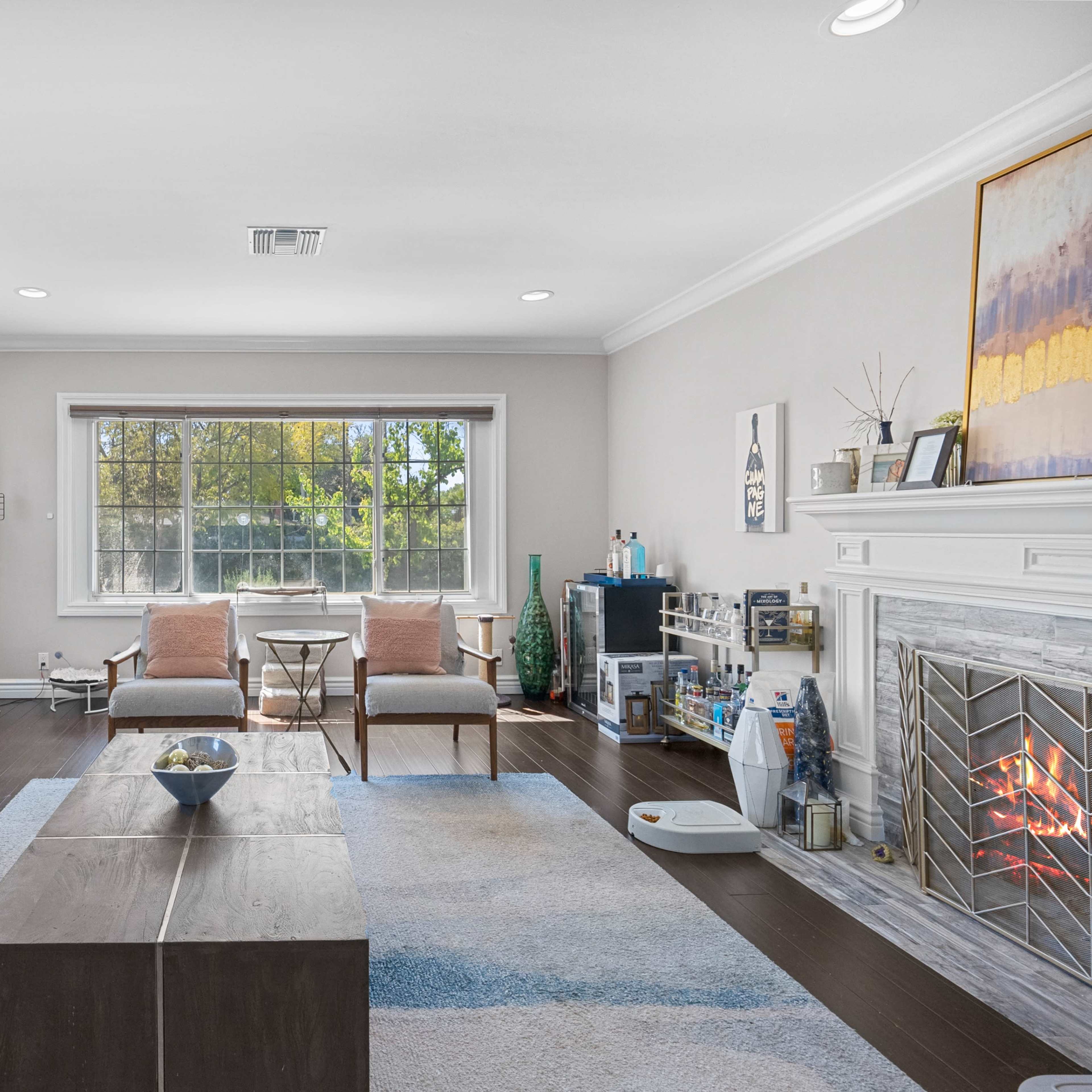 A living room features a blue sofa, two chairs, a coffee table, and a fireplace with a large window providing natural light.