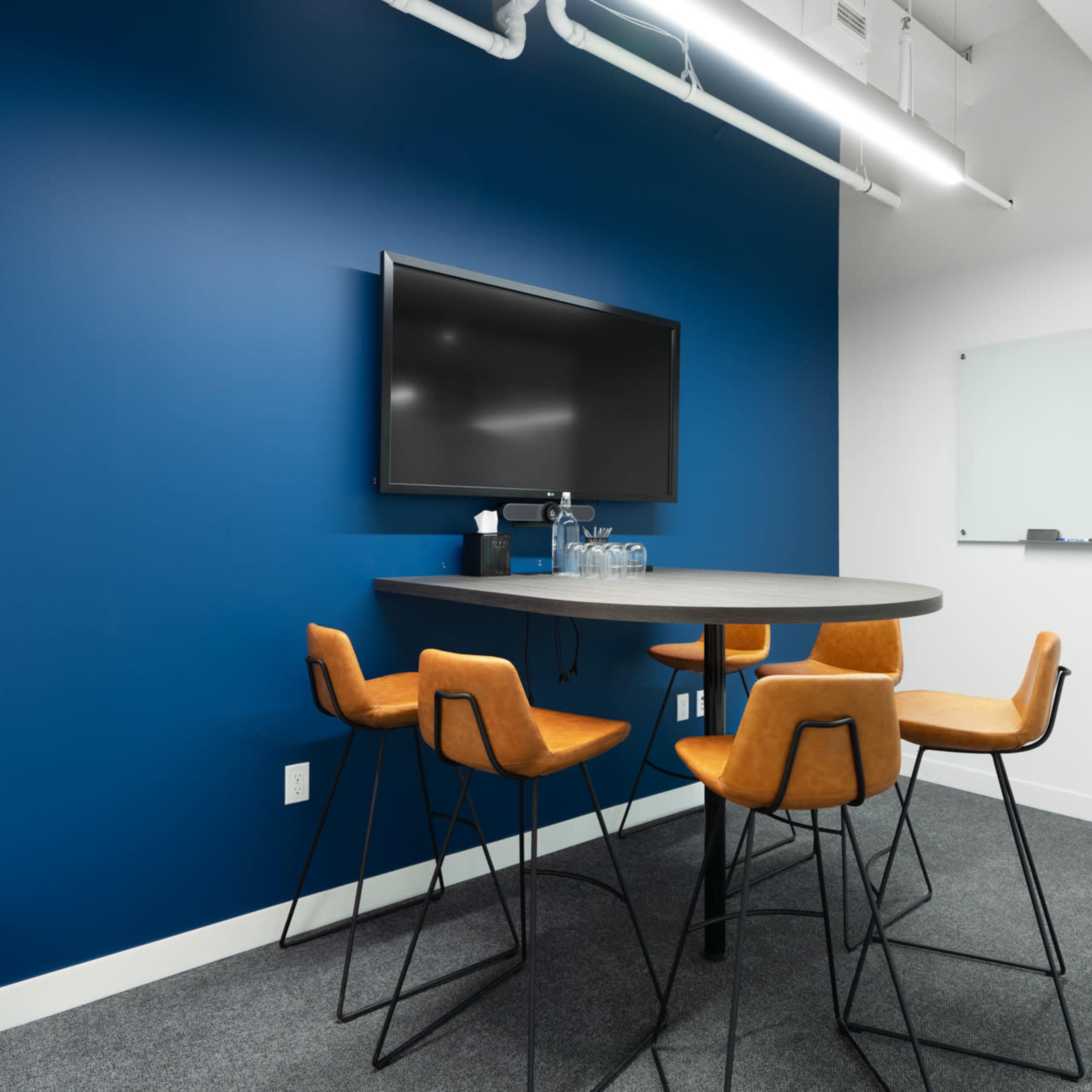A meeting room features a blue wall, a large screen on one side, a round table, and four high-backed chairs around it.