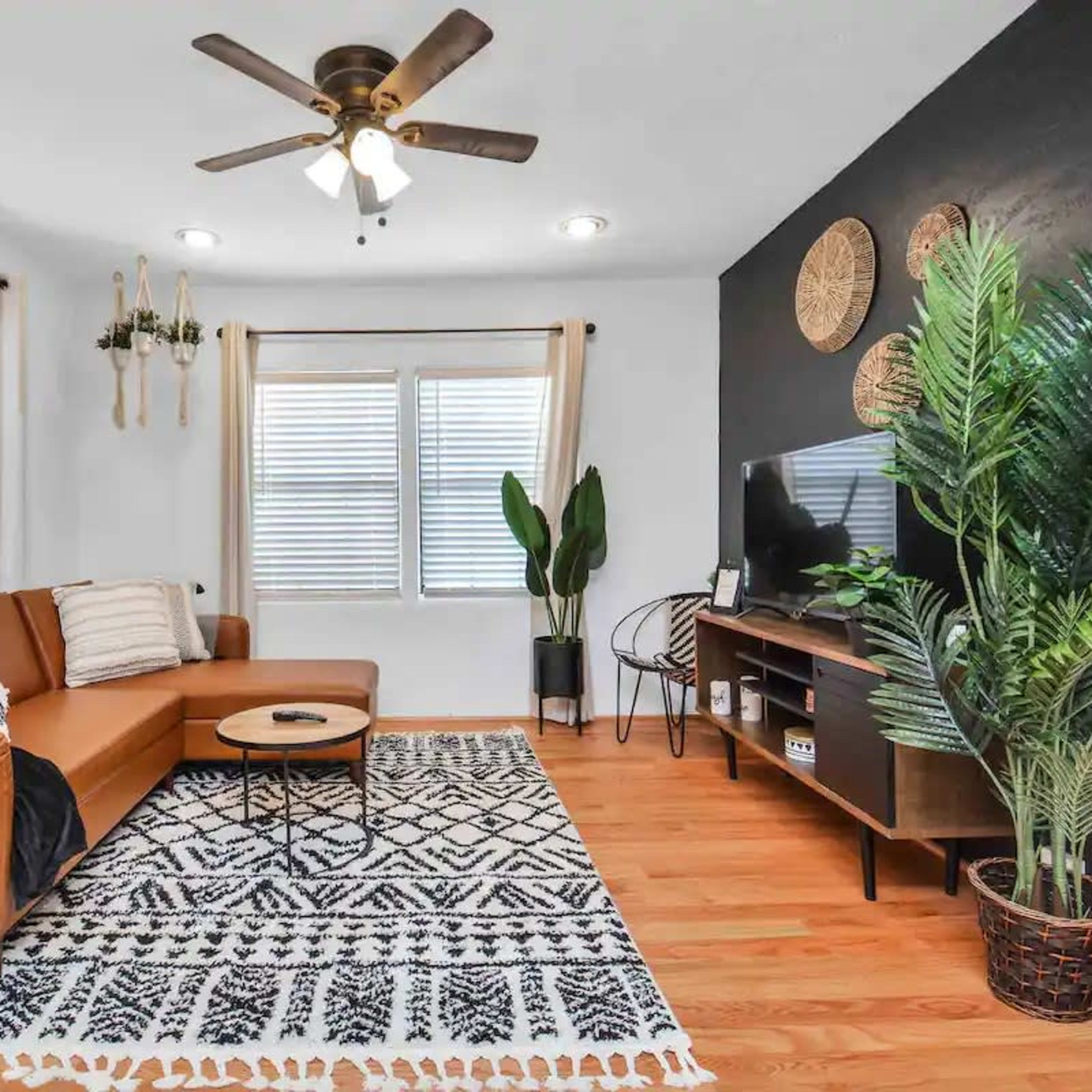 The living room features a brown leather sofa, a round coffee table on a patterned rug, a television mounted on a dark wall, and potted plants.