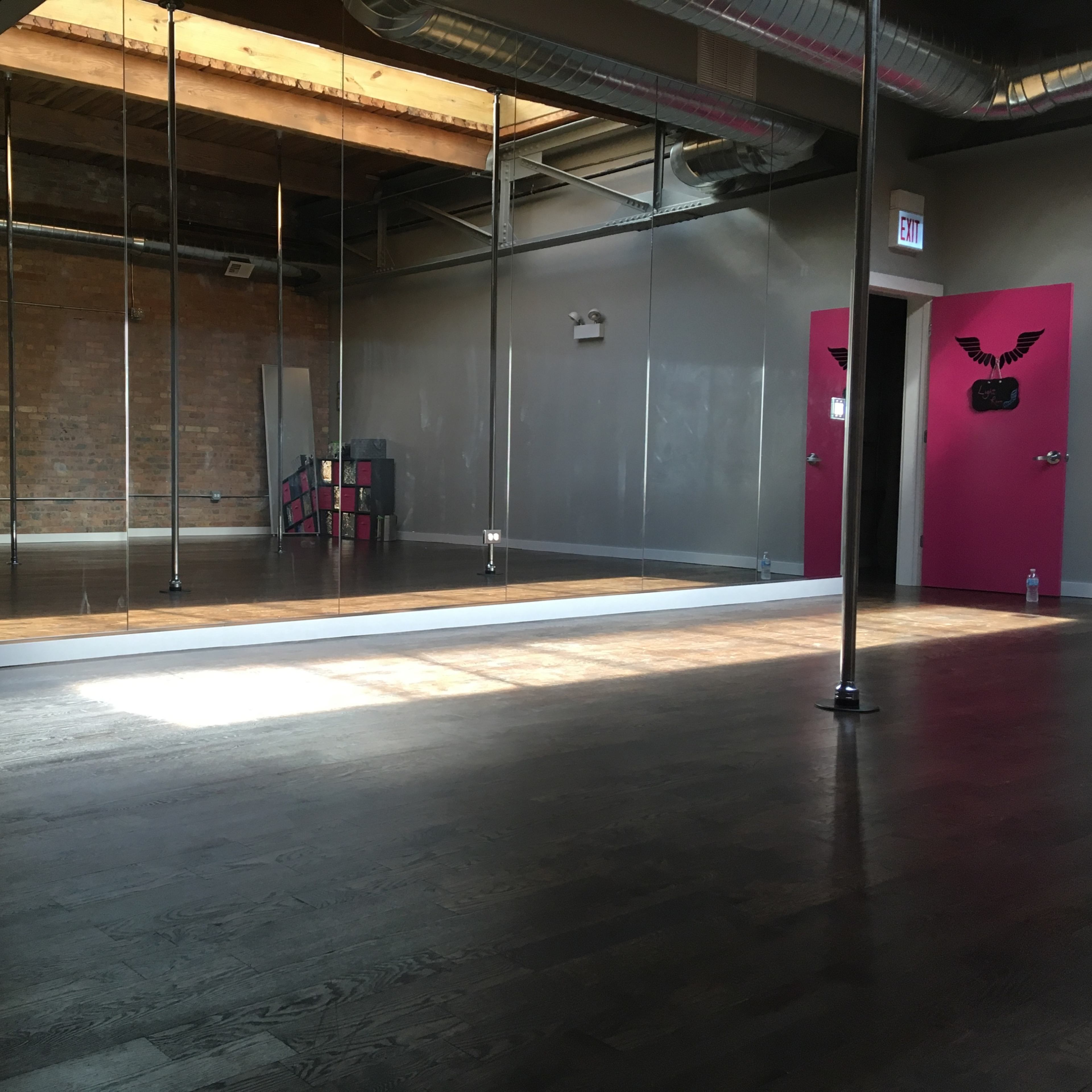 A spacious dance studio with wooden floors, mirrored walls, and a bright pink door at the far end.