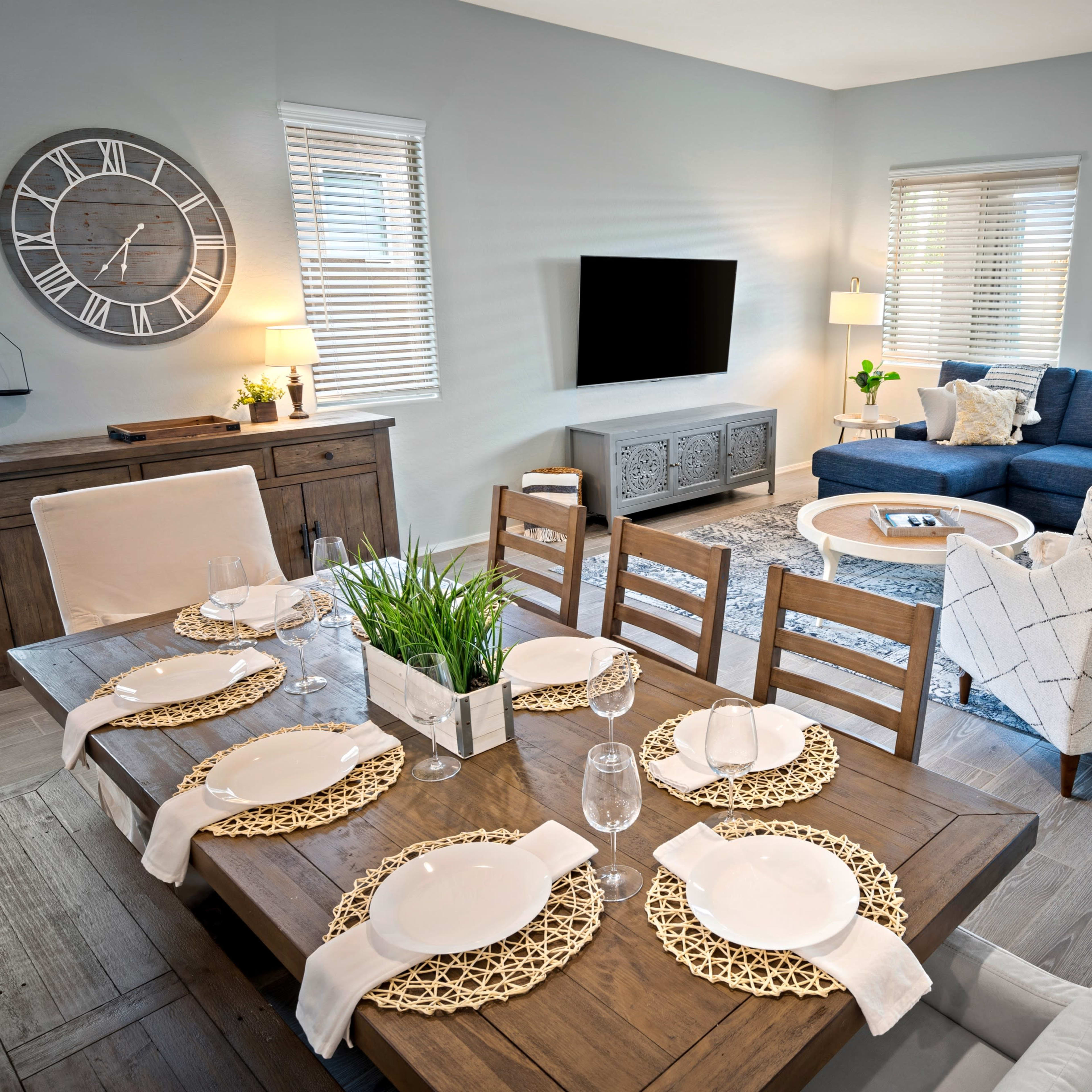 A modern living and dining area featuring a wooden dining table set for six, a dark blue sectional couch, and a wall-mounted TV in a light-colored room with natural light from several windows.