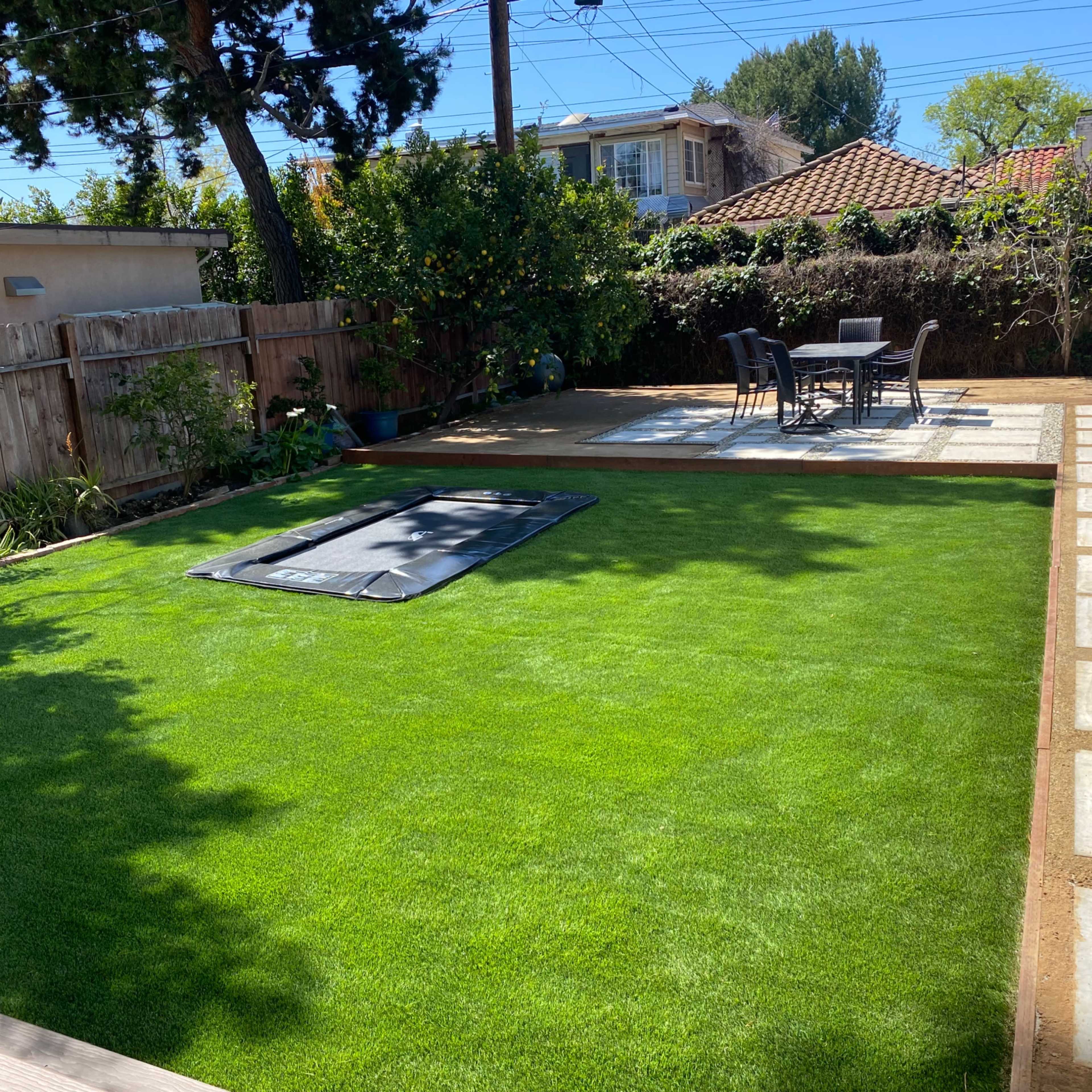A backyard with a green lawn, a rectangular area covered by a tarp, and a patio set with a table and chairs.