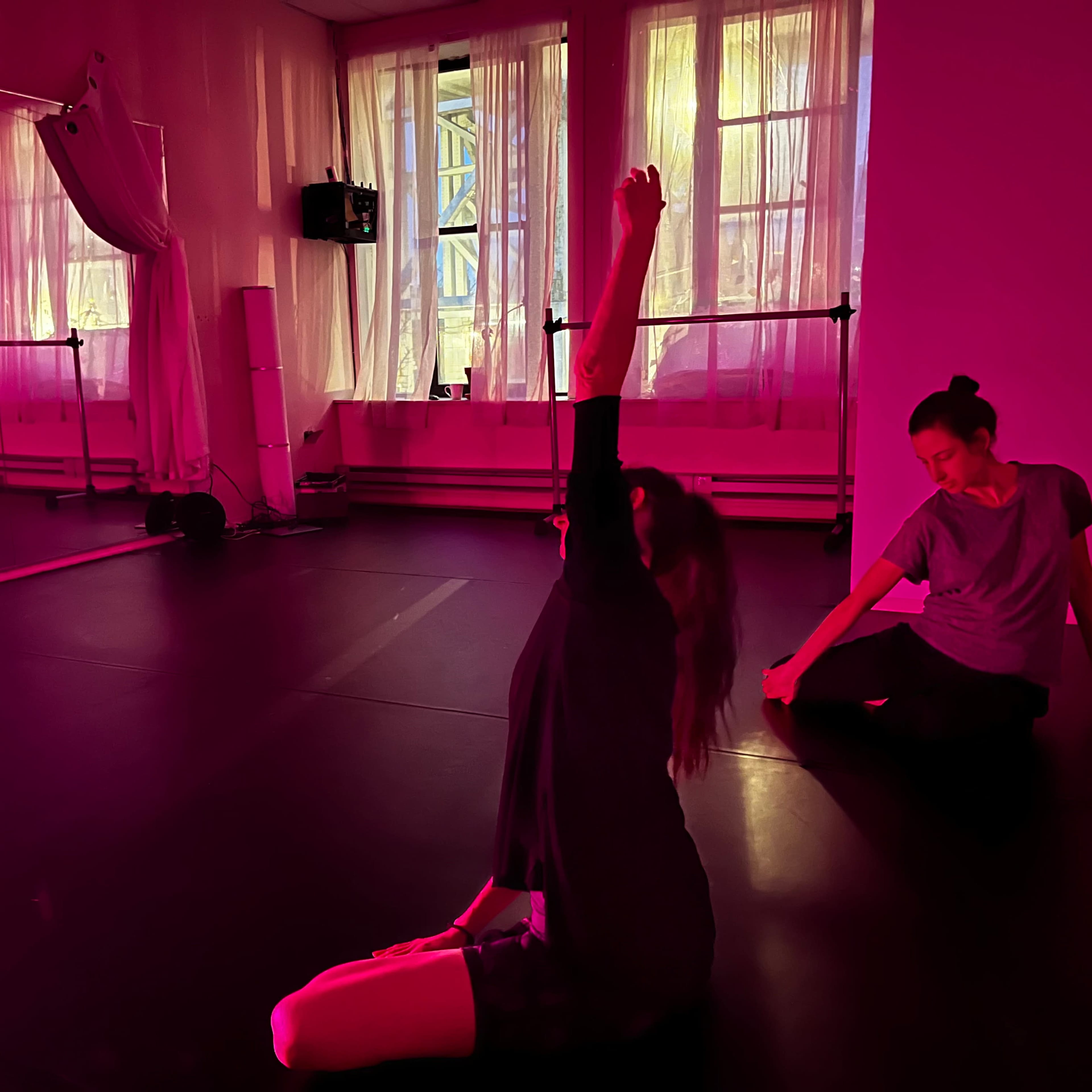 Two dancers practice in a brightly lit studio with pink-tinted lighting, one sitting with an arm raised and the other stretching nearby.