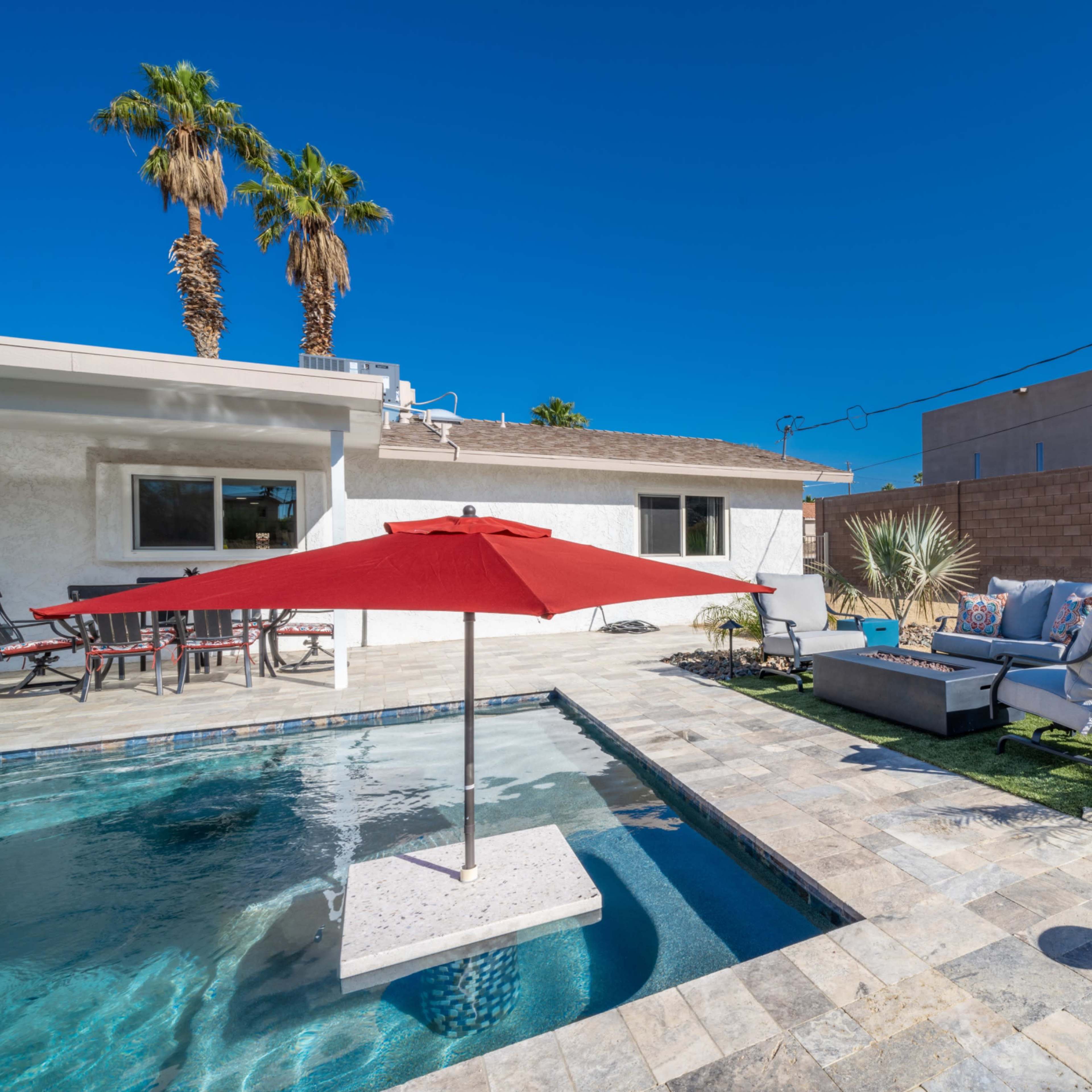 A swimming pool with a red umbrella, surrounded by a paved patio and decorative plants, is situated next to a house.