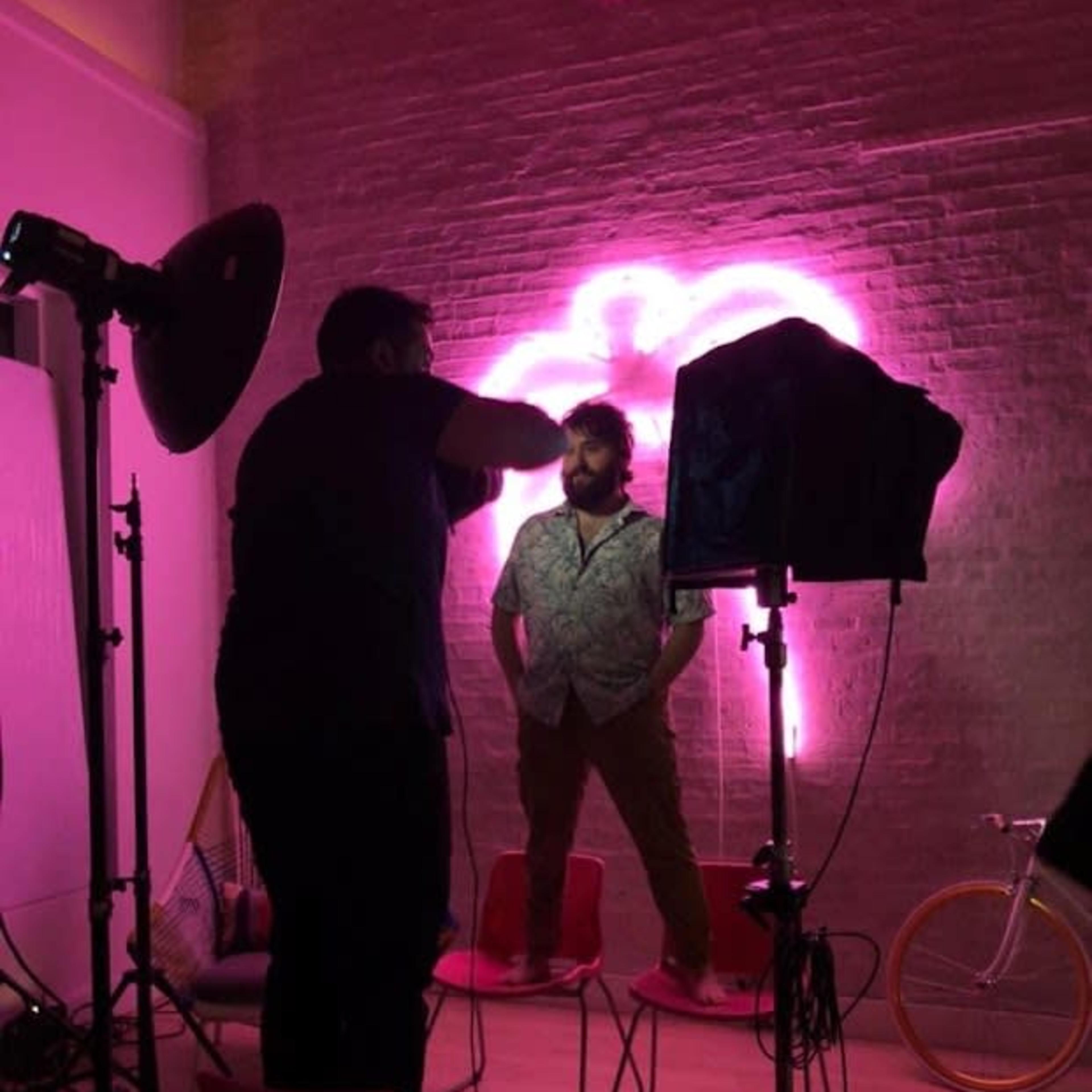 A man stands on chairs in a studio setting, illuminated by a pink neon light, while a photographer adjusts the lighting setup.