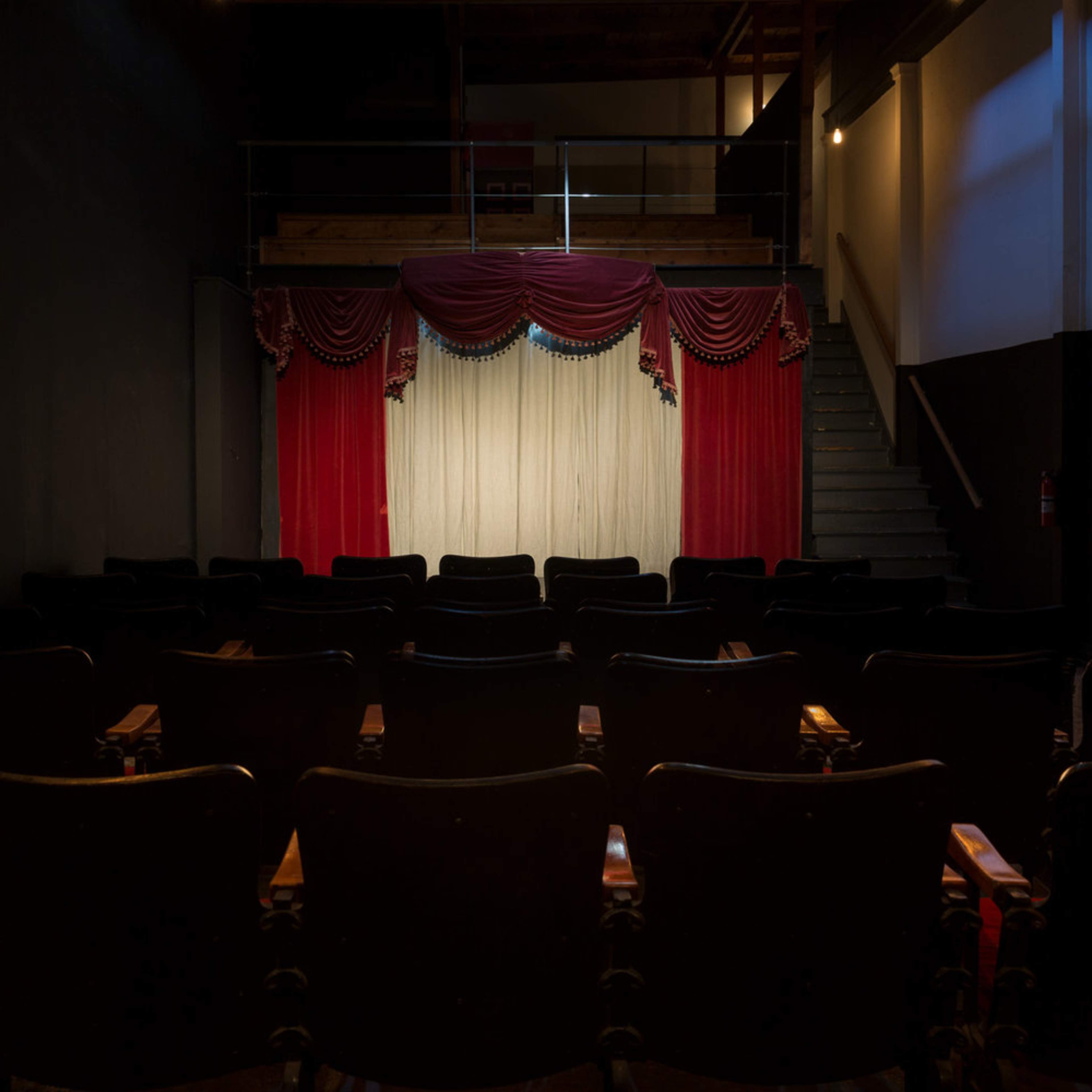 A small theater interior features rows of black audience chairs facing a stage adorned with red curtains and a white backdrop.