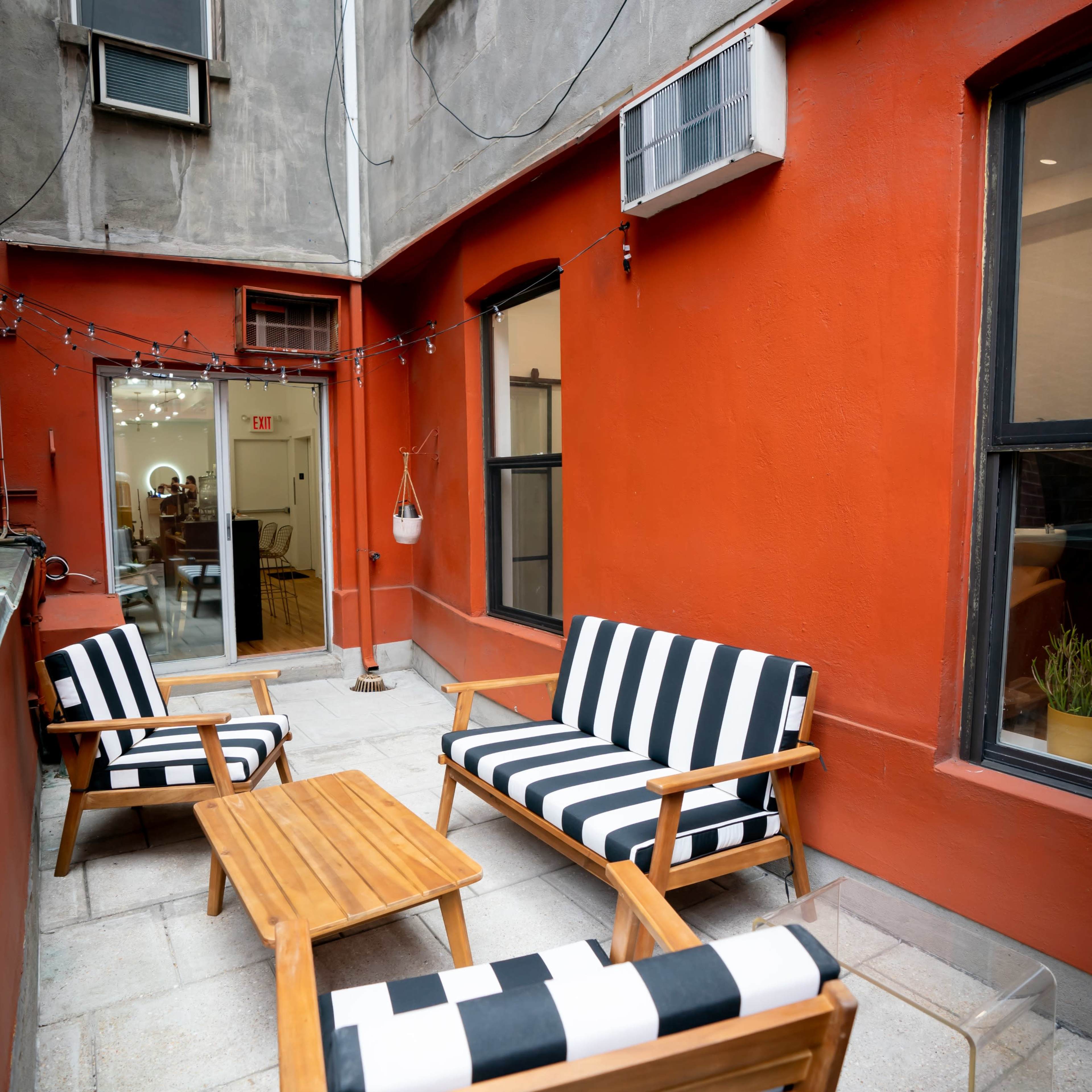 The scene shows a small outdoor patio with striped seating, a wooden table, and an entrance to an interior space, surrounded by orange walls and two air conditioning units.