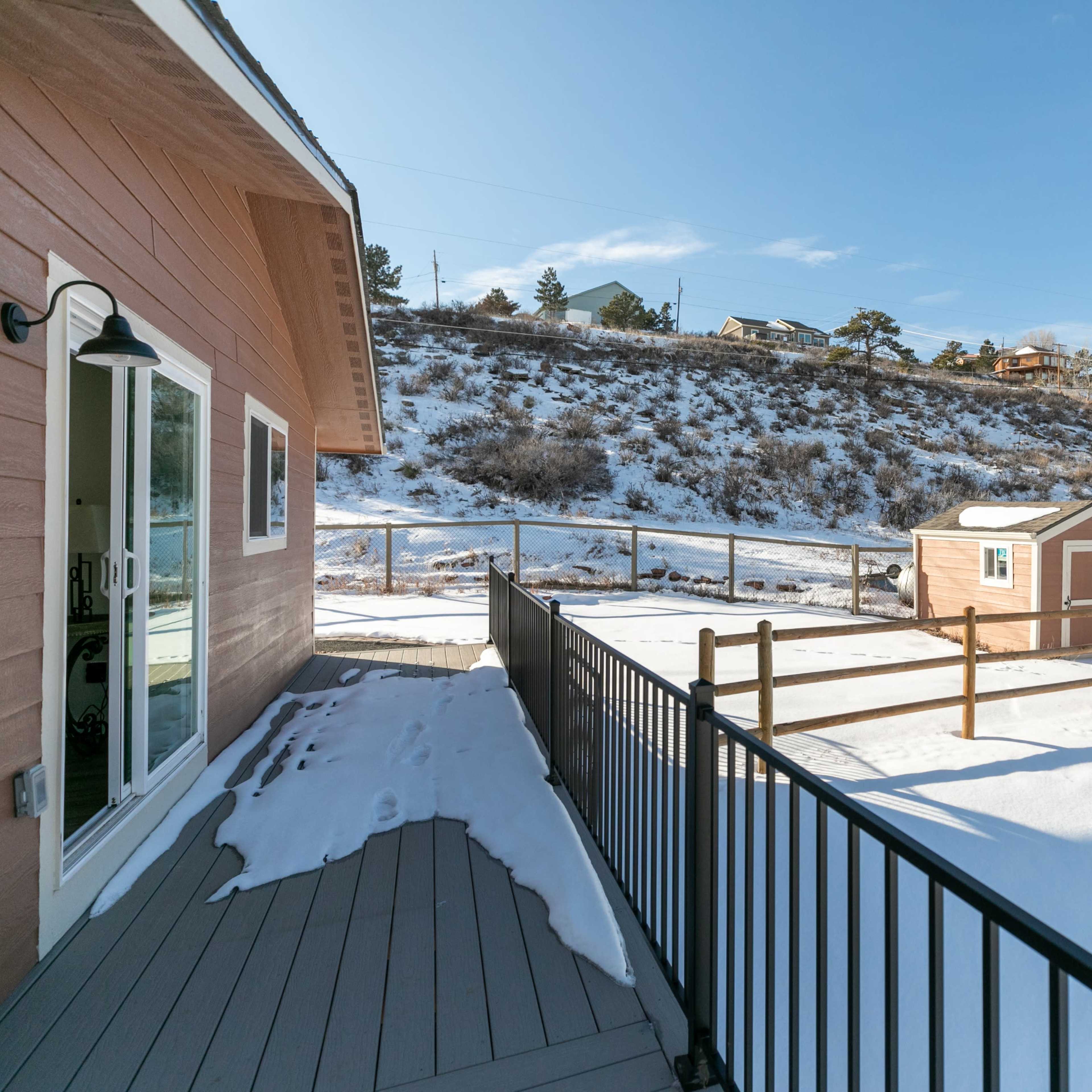 A snow-covered yard with a small shed and a wooden fence against a hillside under a clear blue sky.