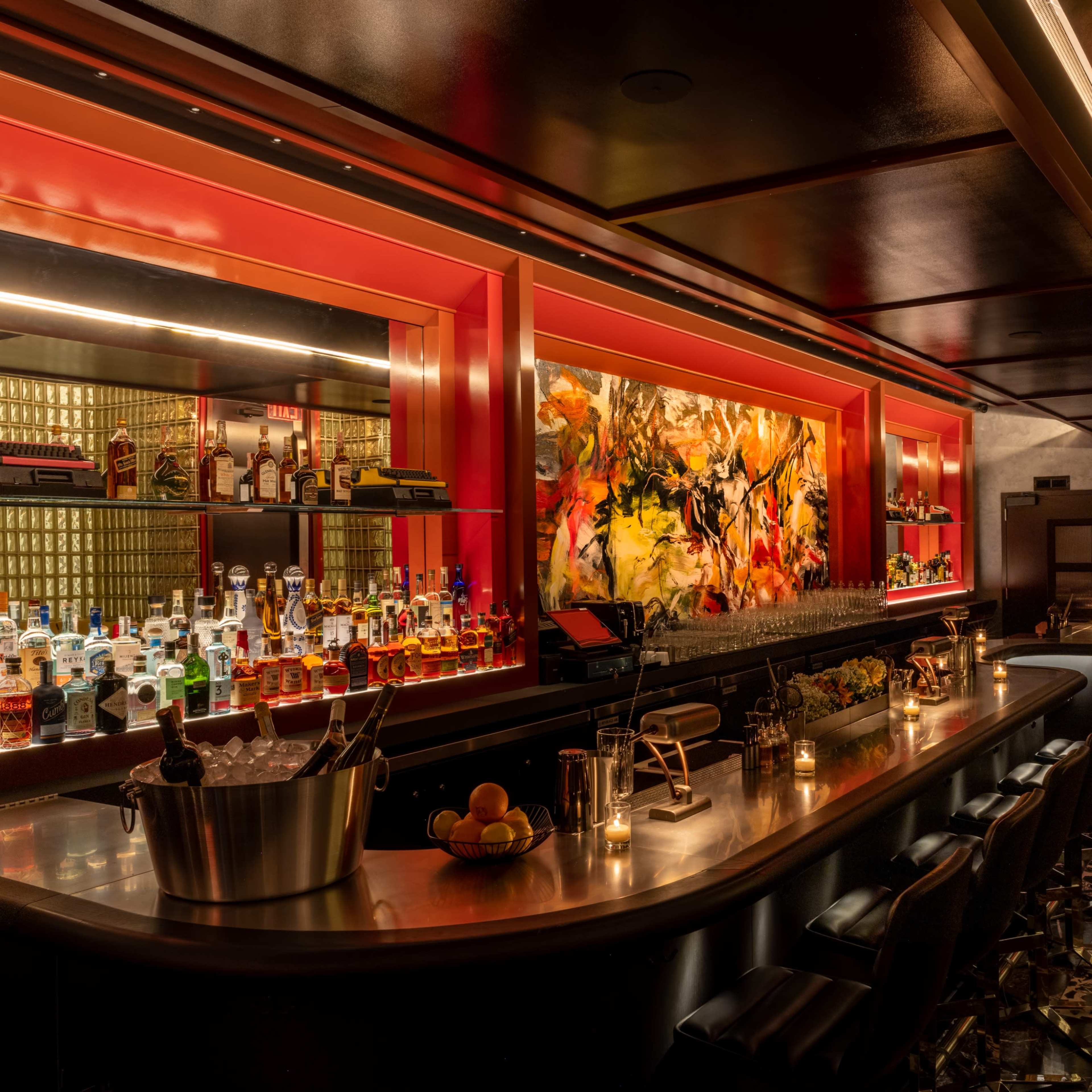 A dimly lit bar with a long countertop, colorful backlit shelves containing a variety of bottles, and a large artwork displayed on the wall.