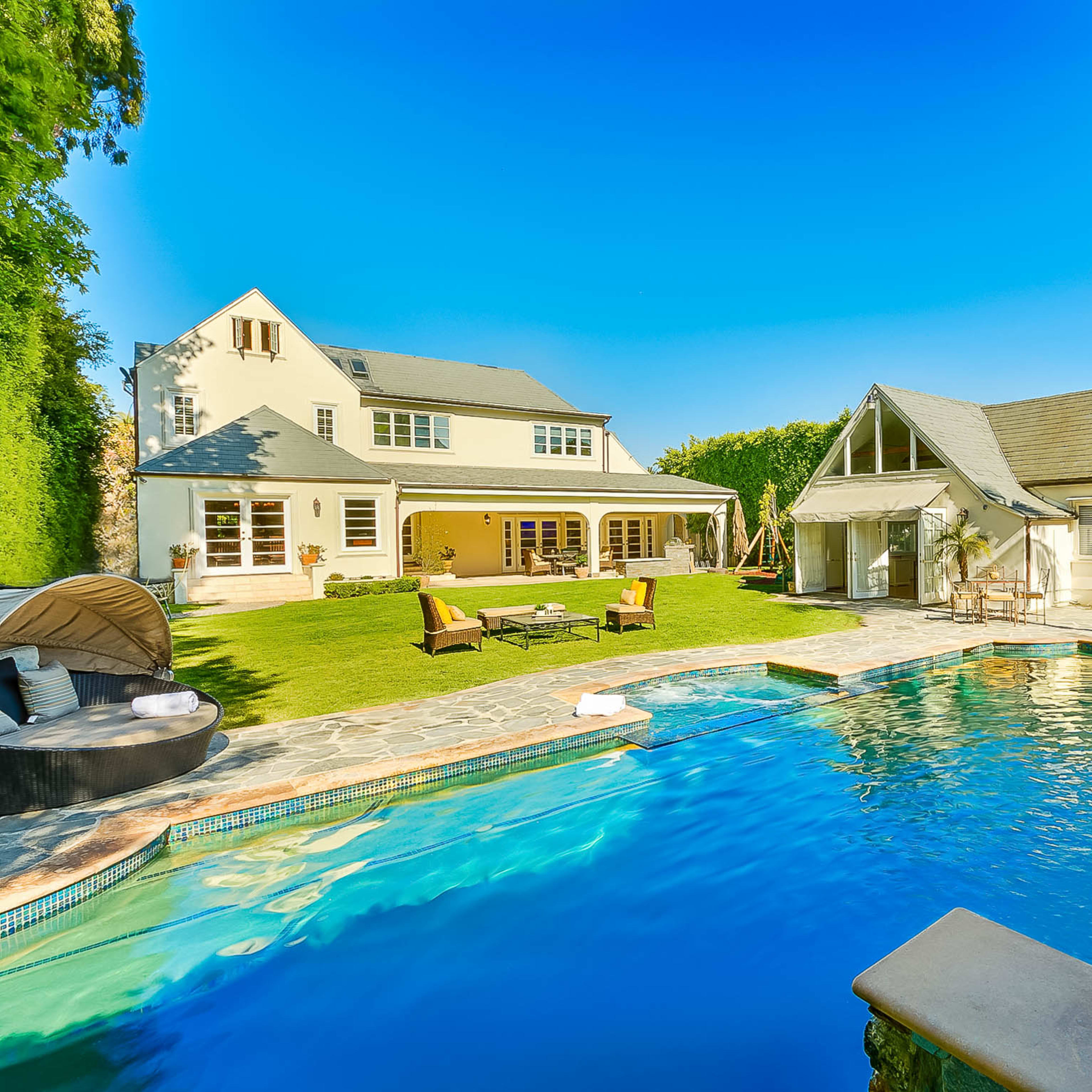 A spacious backyard featuring a swimming pool with a waterfall, surrounded by lounge chairs, a lawn, and two houses in bright sunlight.
