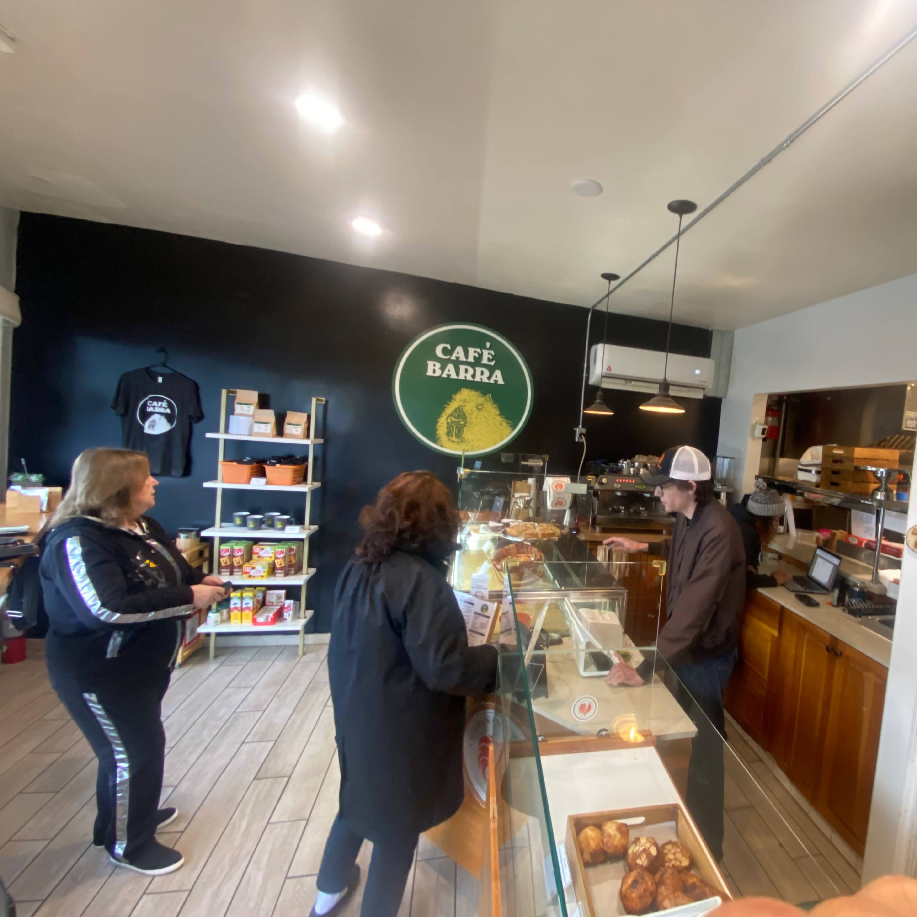 A small café interior shows two customers at the counter while a barista prepares orders behind the glass display.