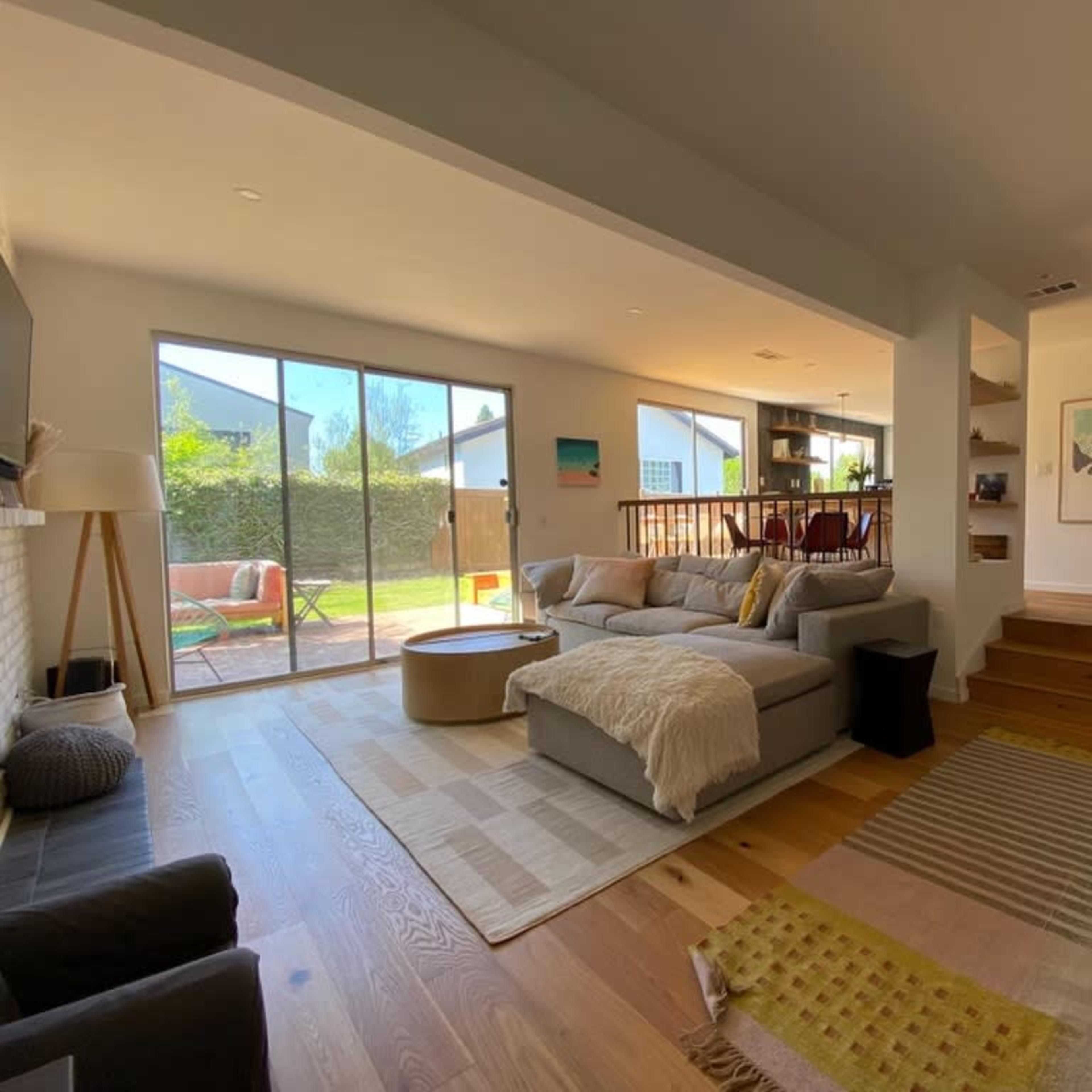A modern living room with a large sectional sofa, a coffee table, and large windows overlooking a backyard, with a staircase leading to an upper level.