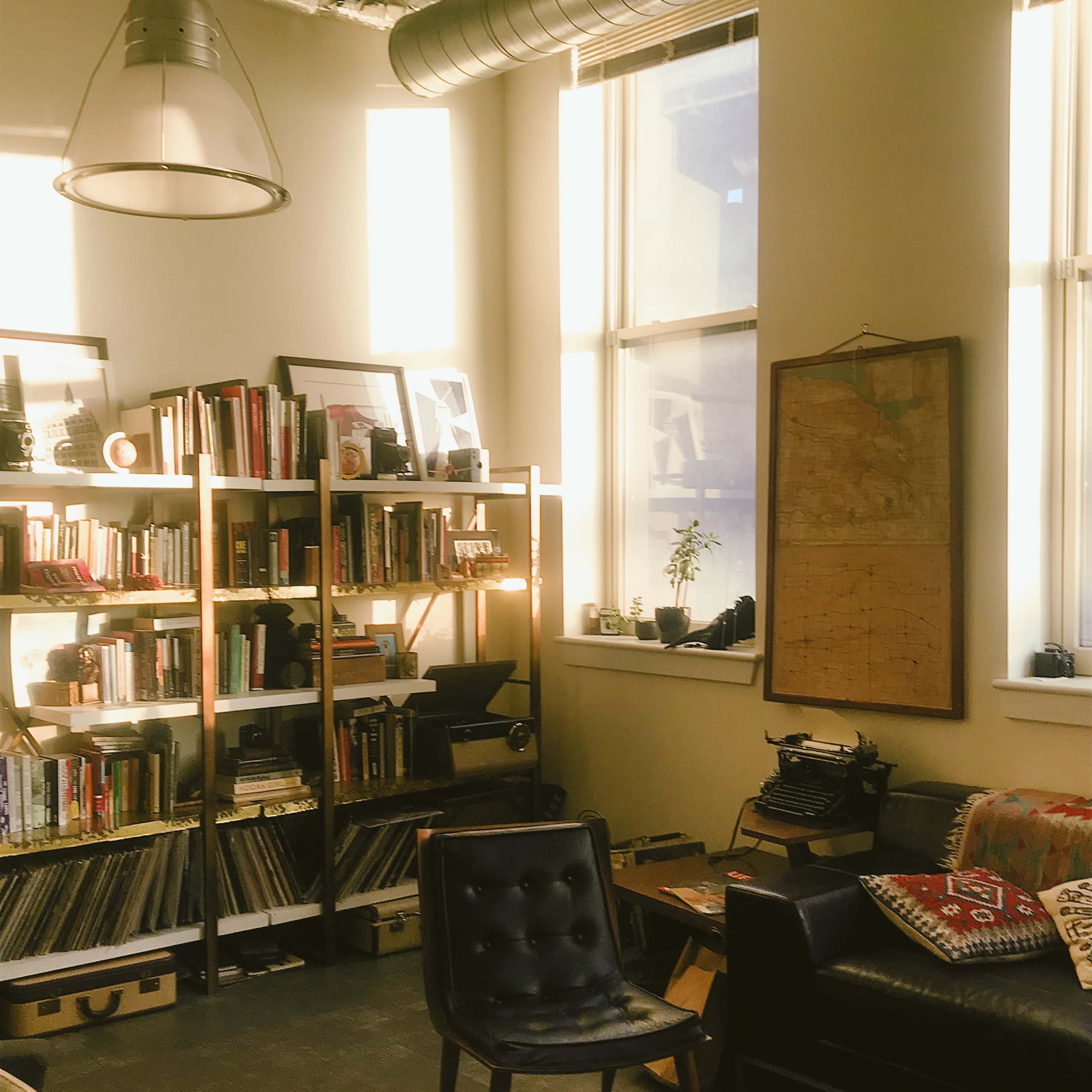 A cozy reading nook featuring a bookshelf filled with books, a vintage typewriter, and a comfortable seating area illuminated by natural light from large windows.