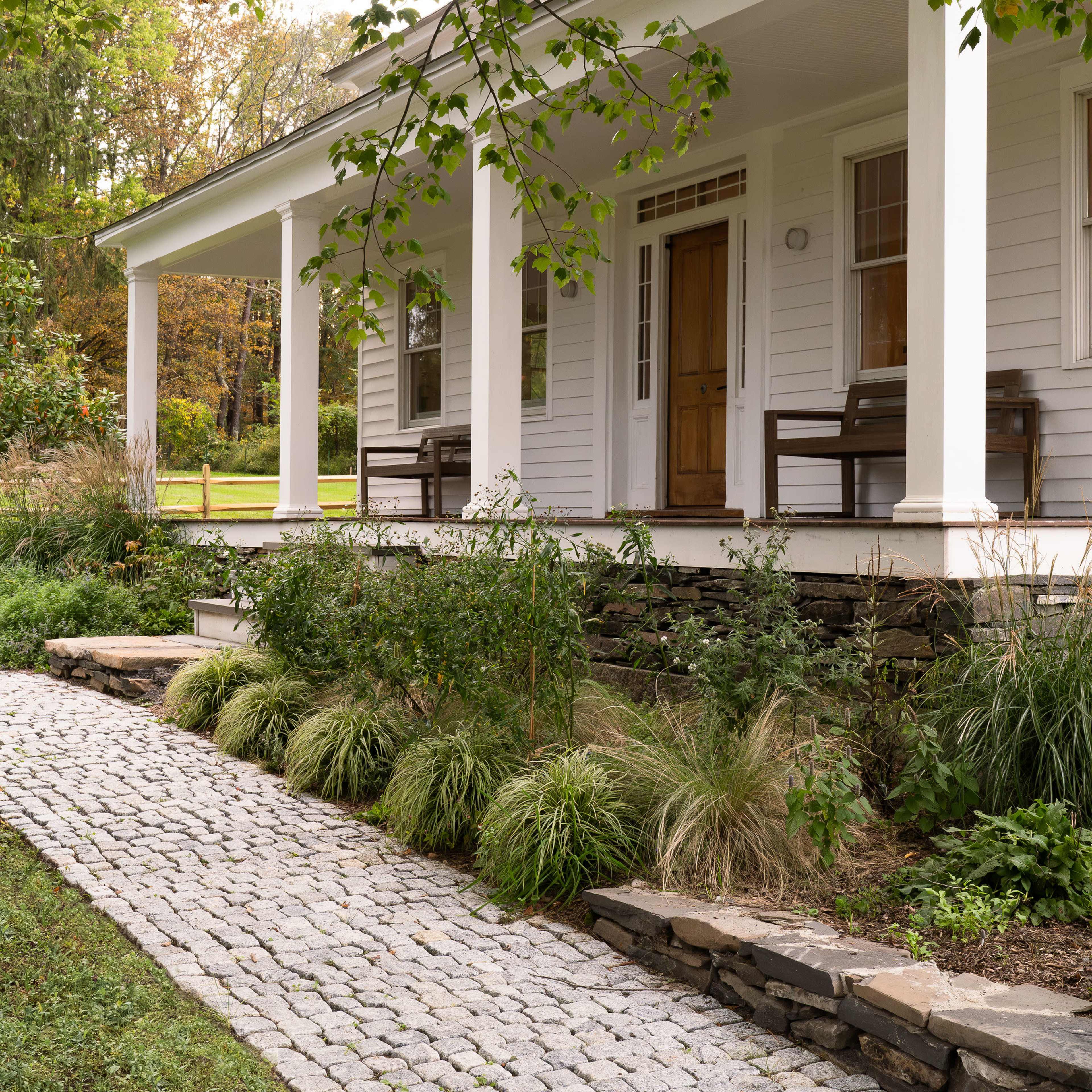 A white house with a front porch is flanked by two benches and surrounded by landscaped greenery, leading to a stone pathway.
