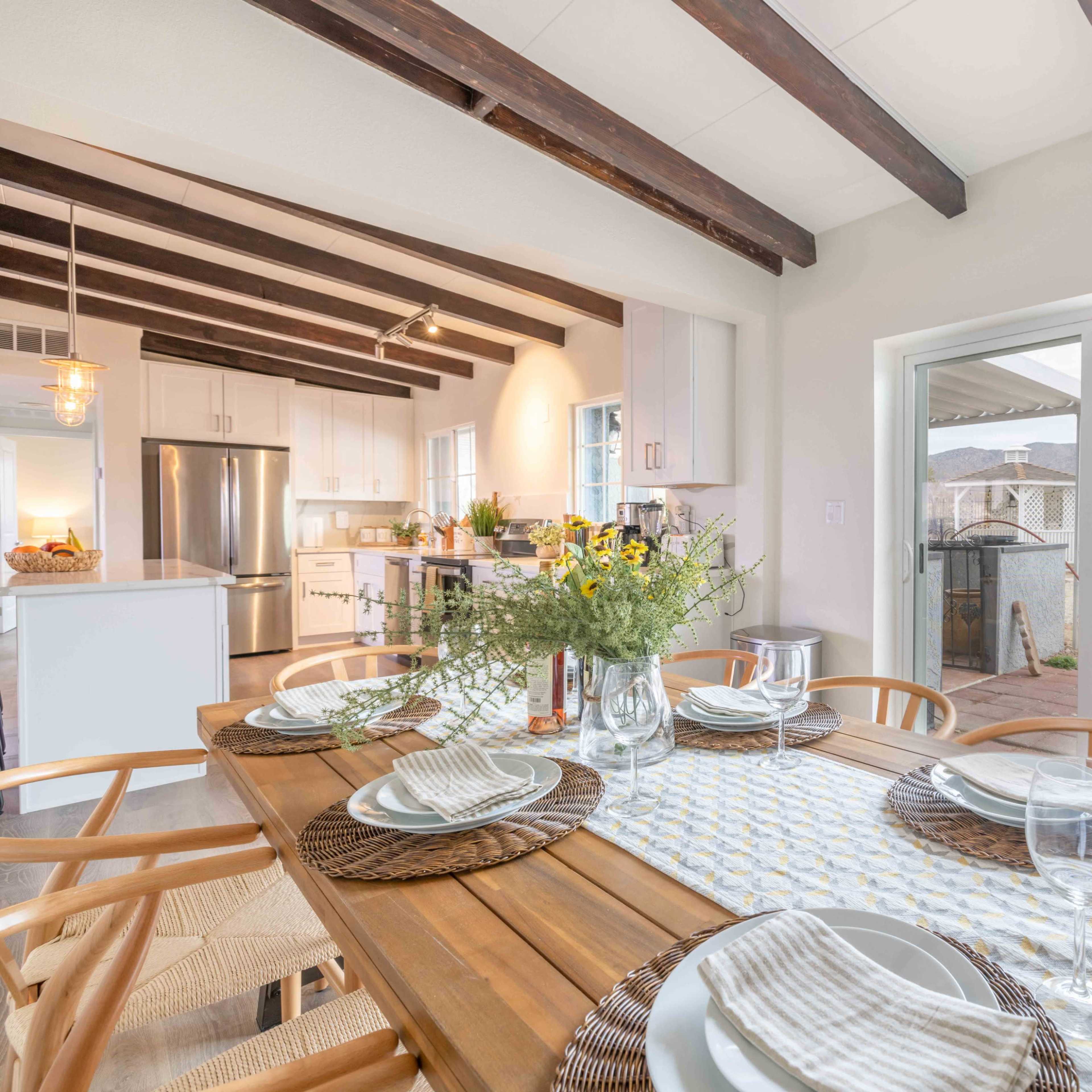 A dining table is set for a meal in a kitchen that opens to a outdoor view with mountains in the background.