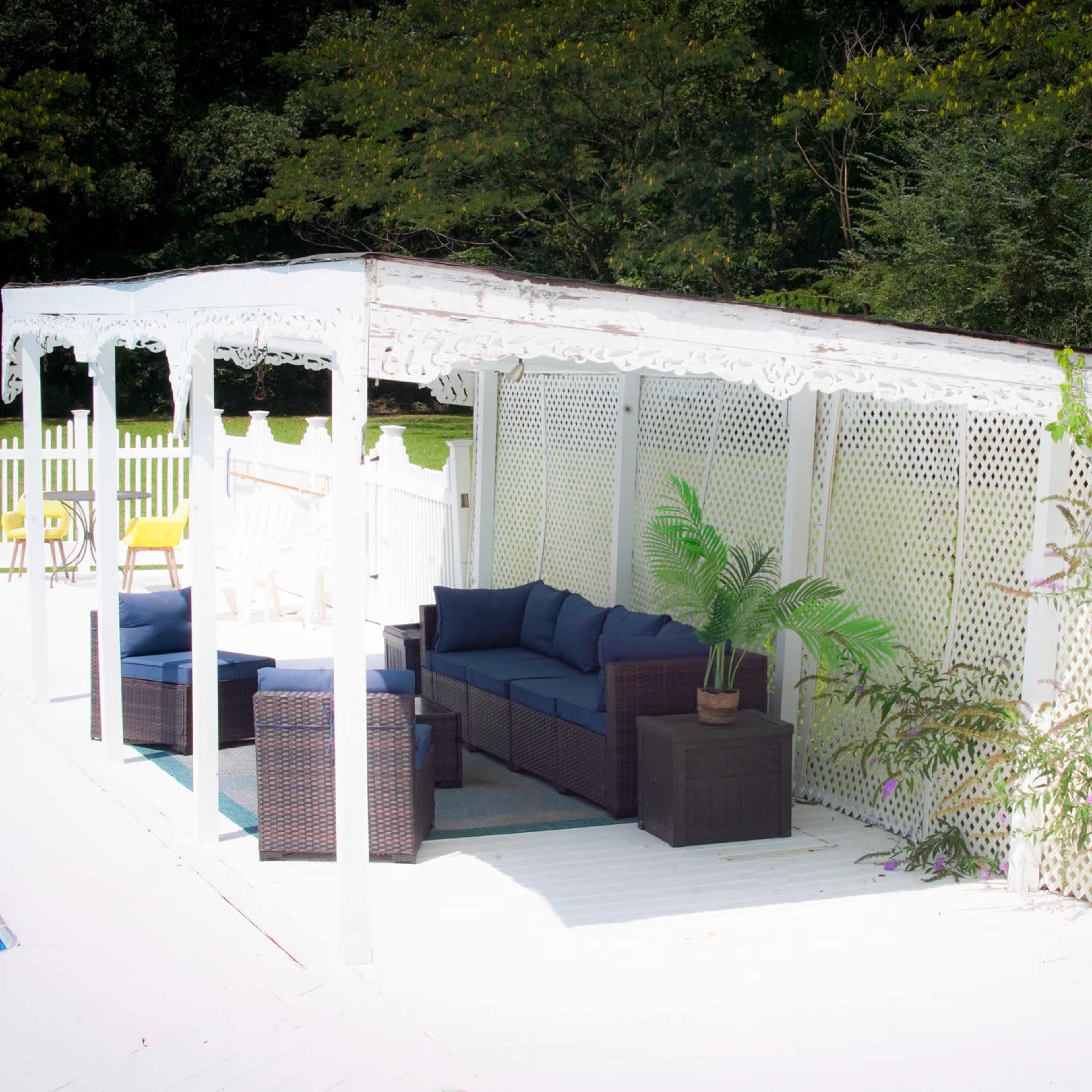 A white outdoor pergola with a seating area beside a swimming pool, surrounded by a wooden fence and greenery.