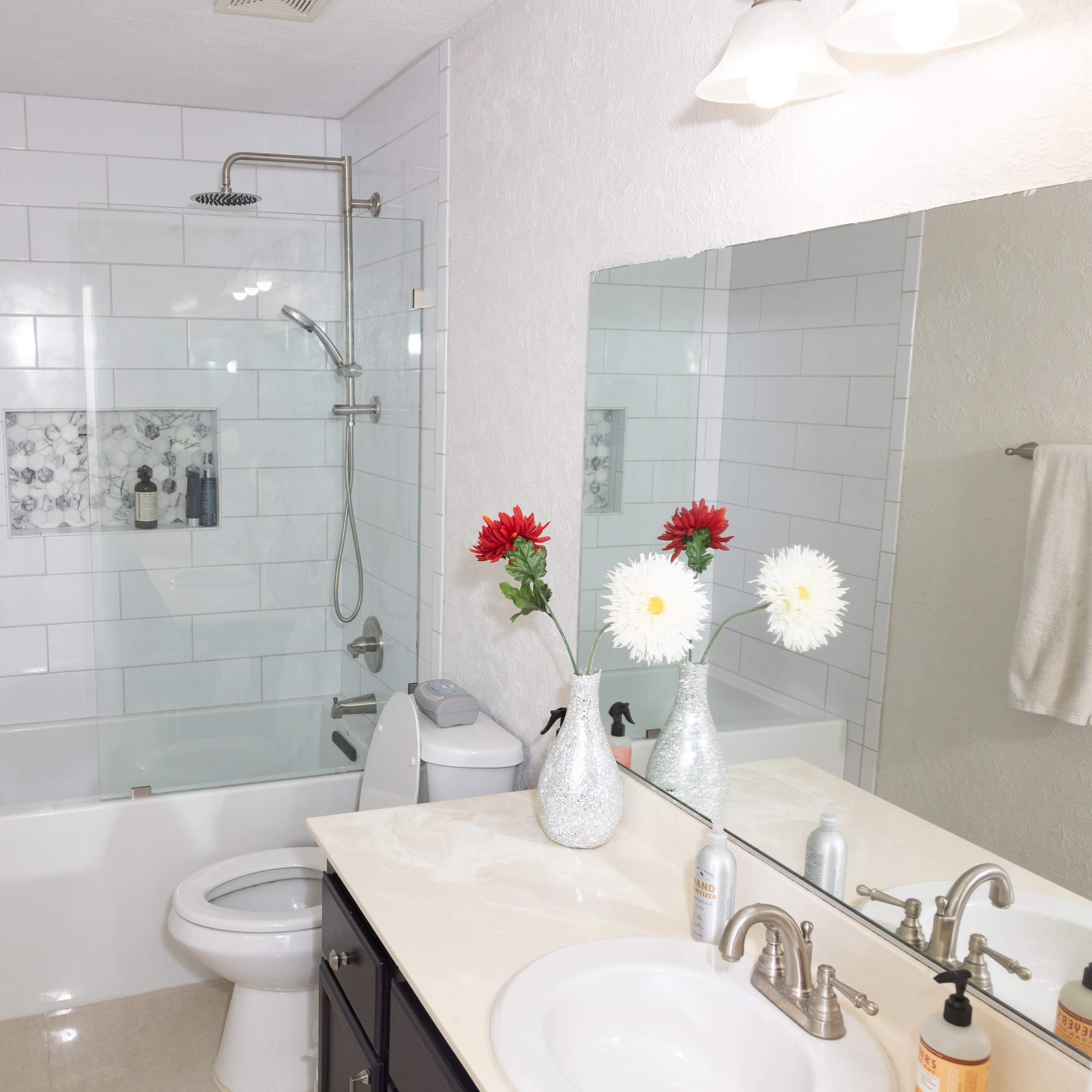 The bathroom features a glass shower enclosure, a bathtub, a toilet, and a countertop with two vases of flowers and various toiletries.