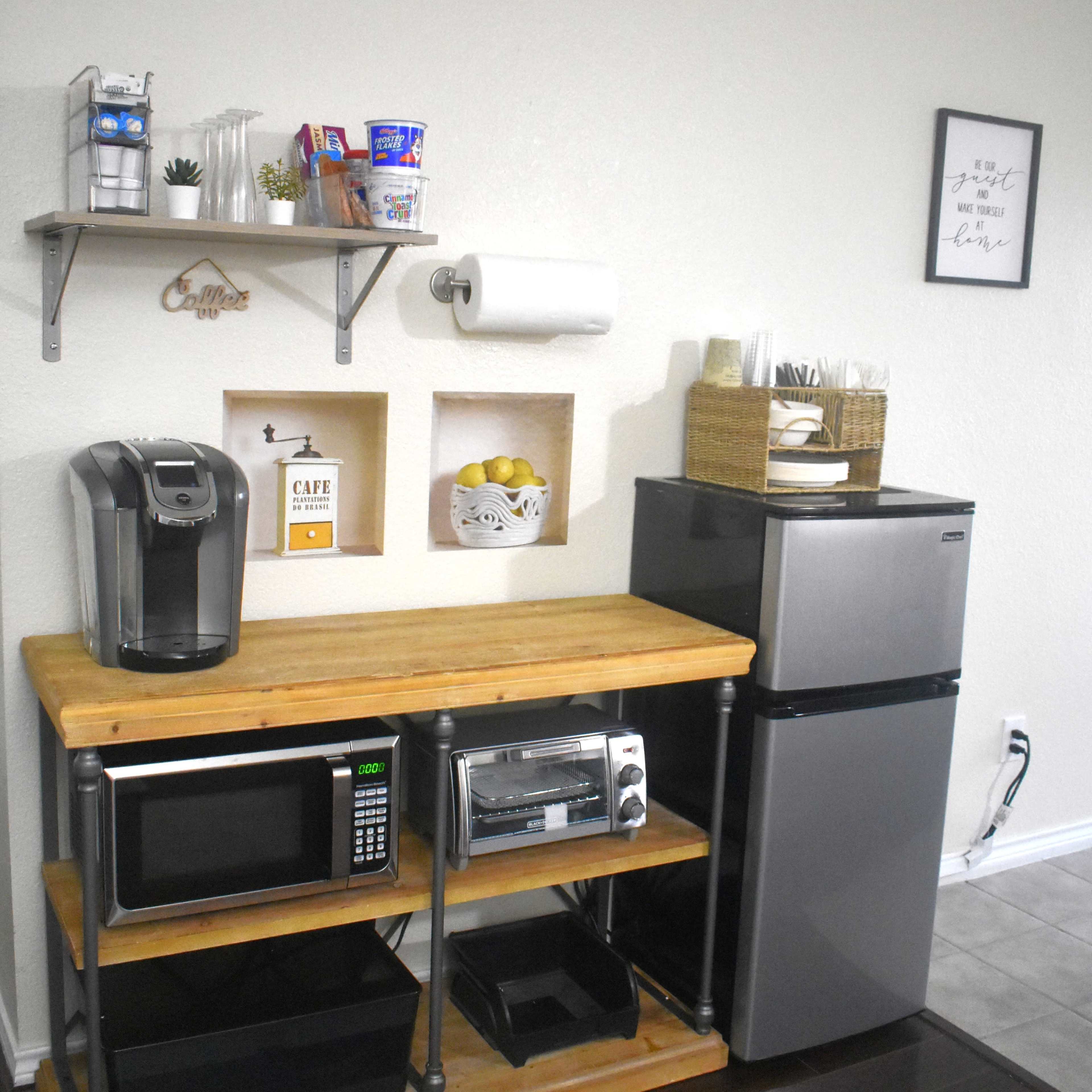 A small kitchen area with a wooden countertop, a coffee maker, a microwave, a toaster oven, a mini fridge, and shelves displaying snacks and kitchen supplies.