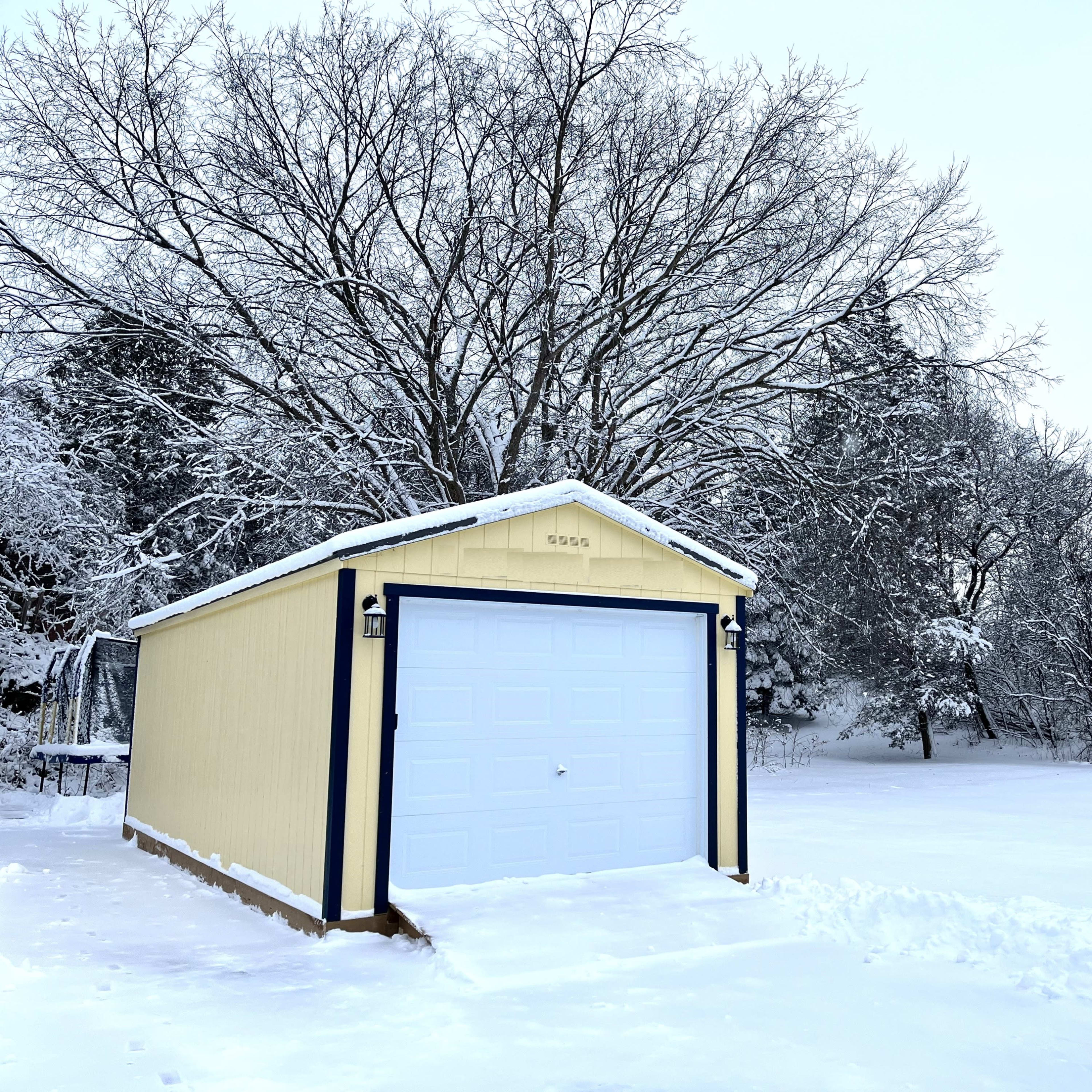 A yellow shed with a white garage door is surrounded by a snowy landscape and bare trees.