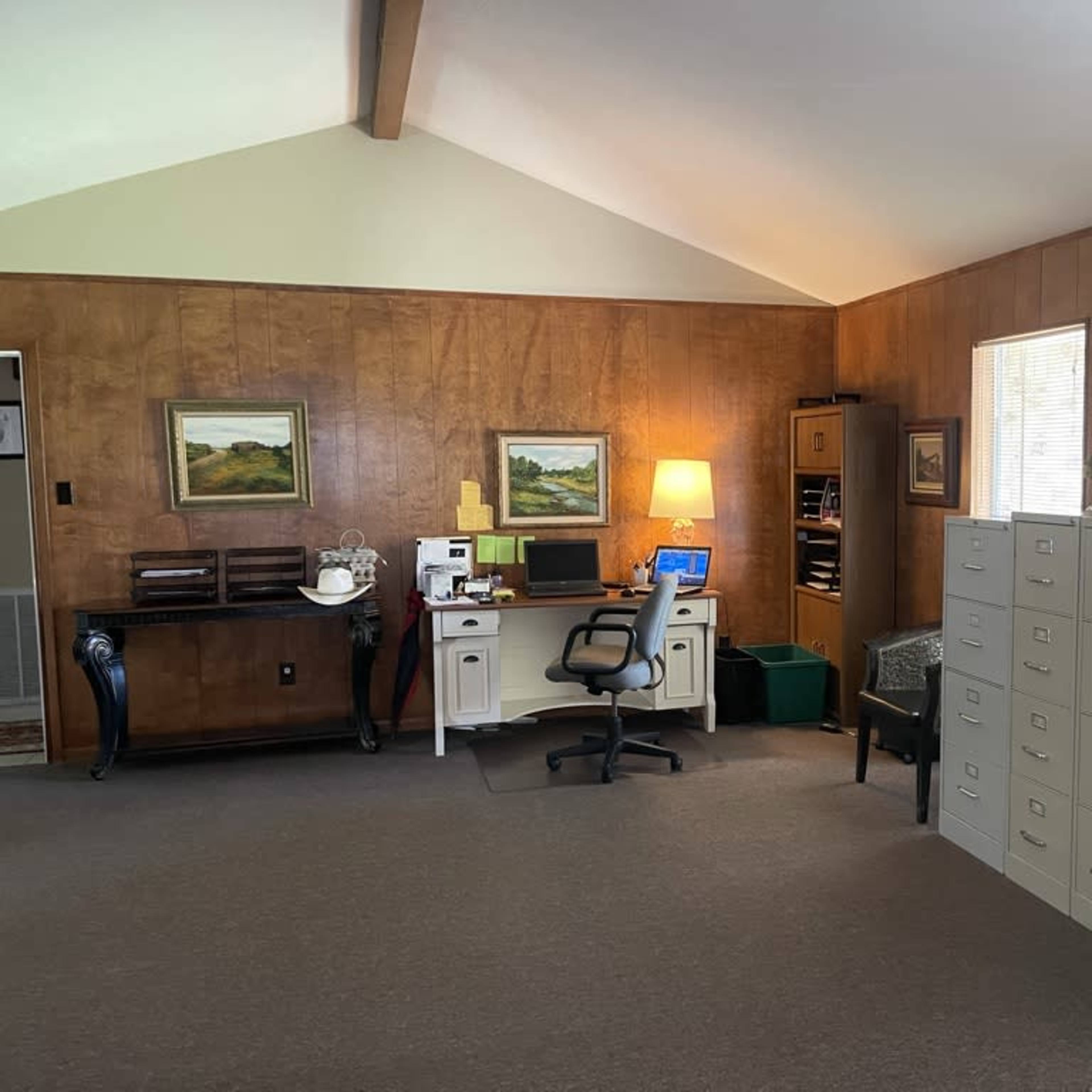 An office space with wooden walls, a desk with a computer and chair, filing cabinets, and soft lighting from a lamp.