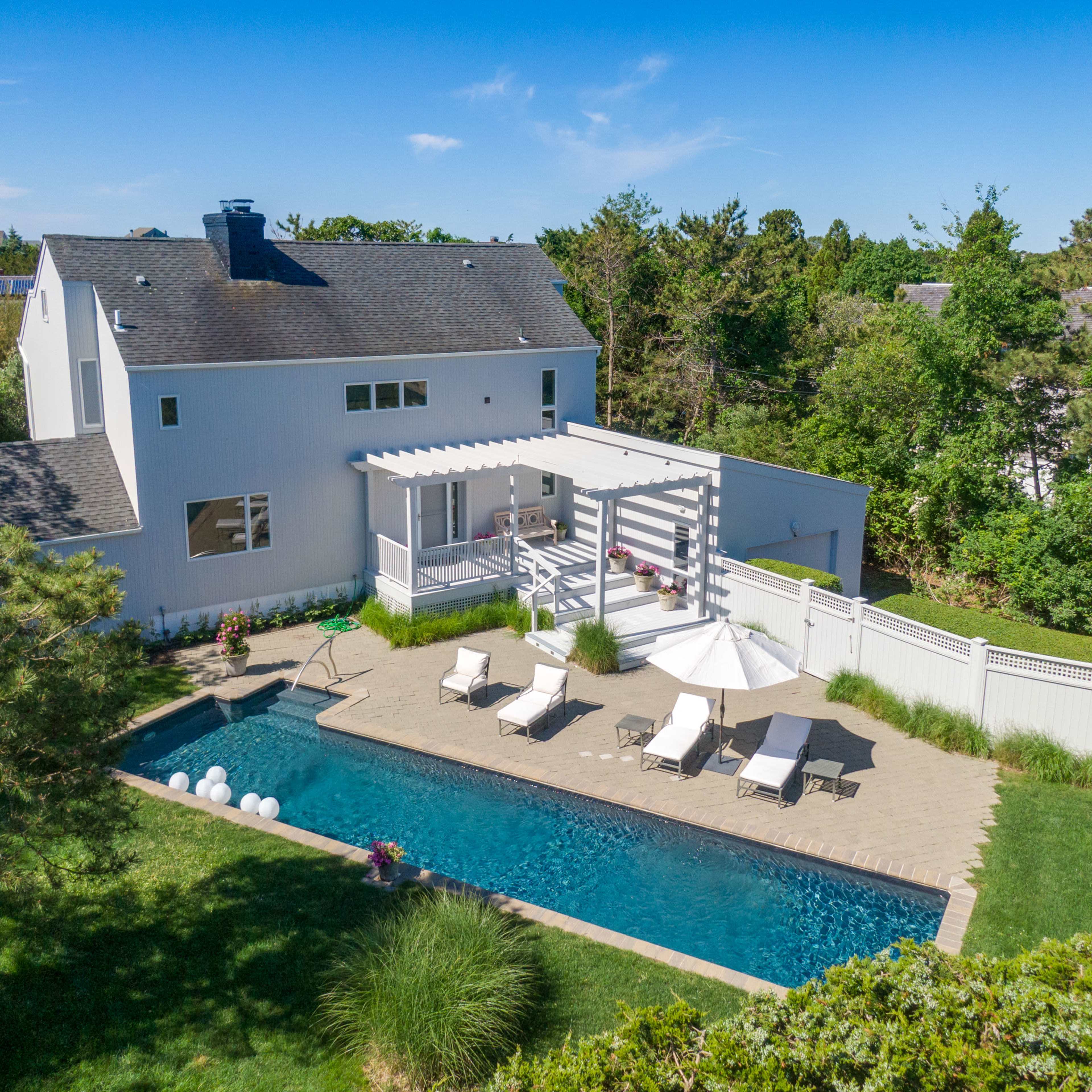 A spacious backyard features a rectangular pool surrounded by lounge chairs and a patio area, with a house and trees in the background.