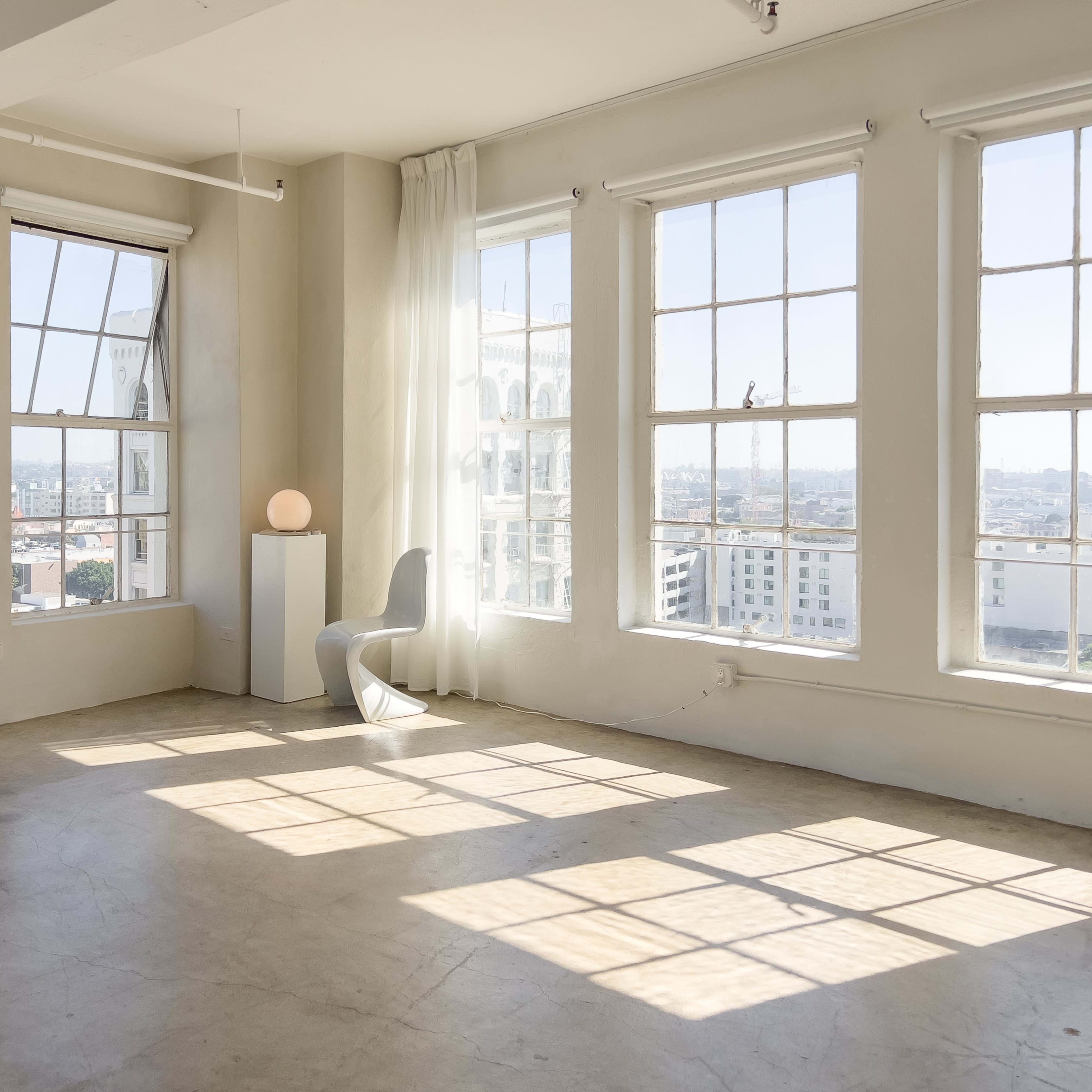 A bright, minimalistic room with large windows, a sculptural chair, and a small lamp on a pedestal.