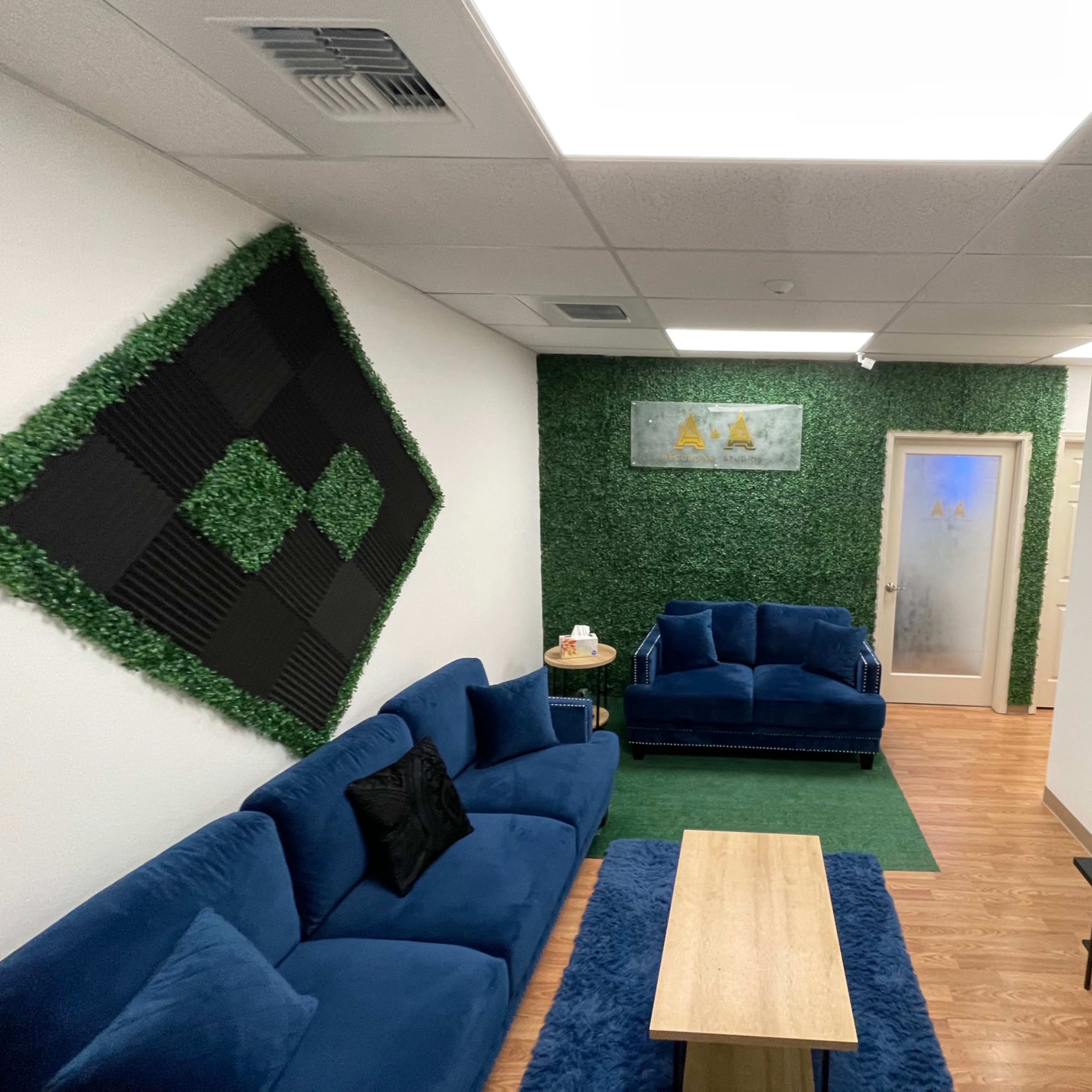 A modern lounge area with blue sofas, a wooden coffee table, artificial greenery on the walls, and a television mounted on one side.