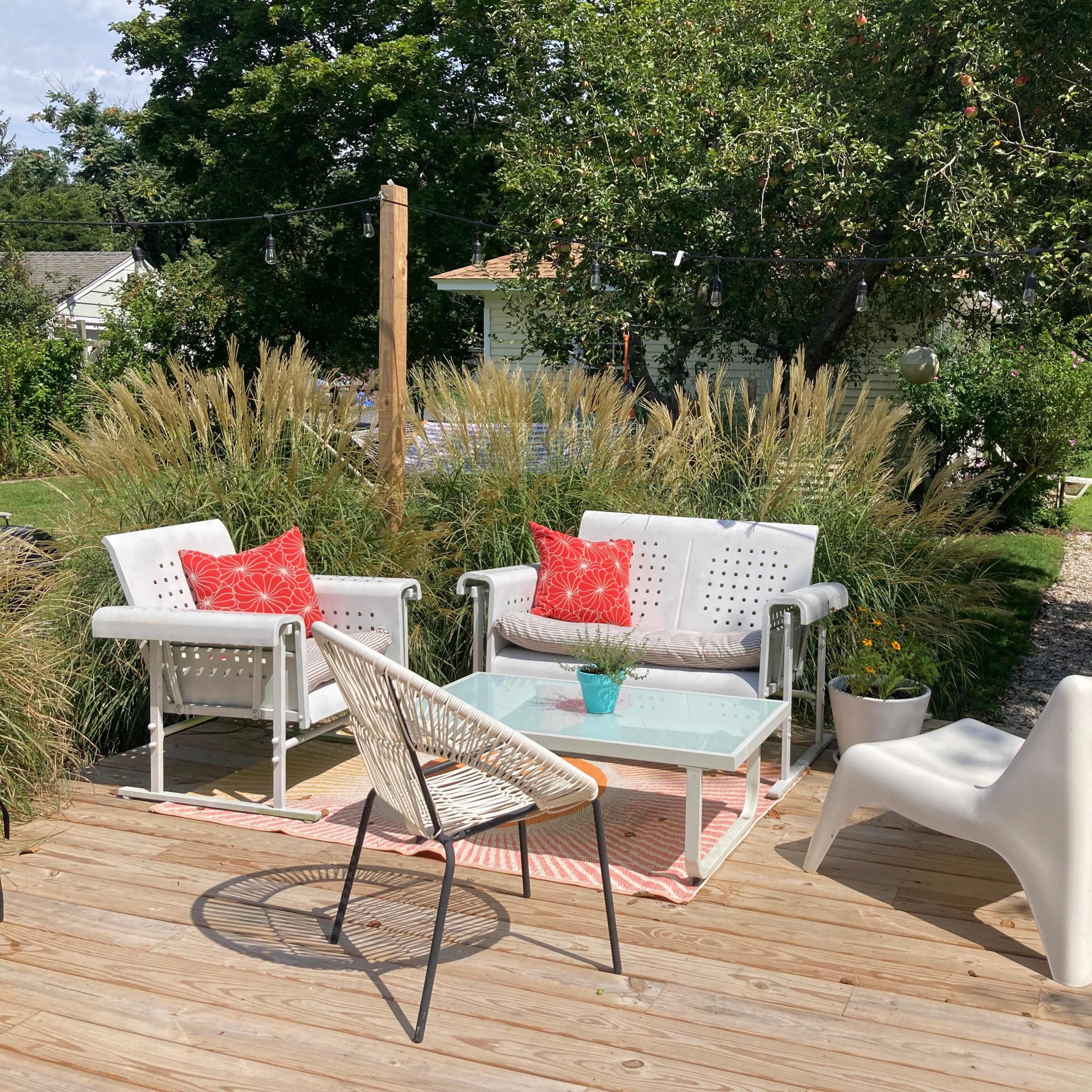 A backyard scene with a wooden deck featuring white outdoor seating, a glass coffee table, and ornamental grasses surrounding the area.