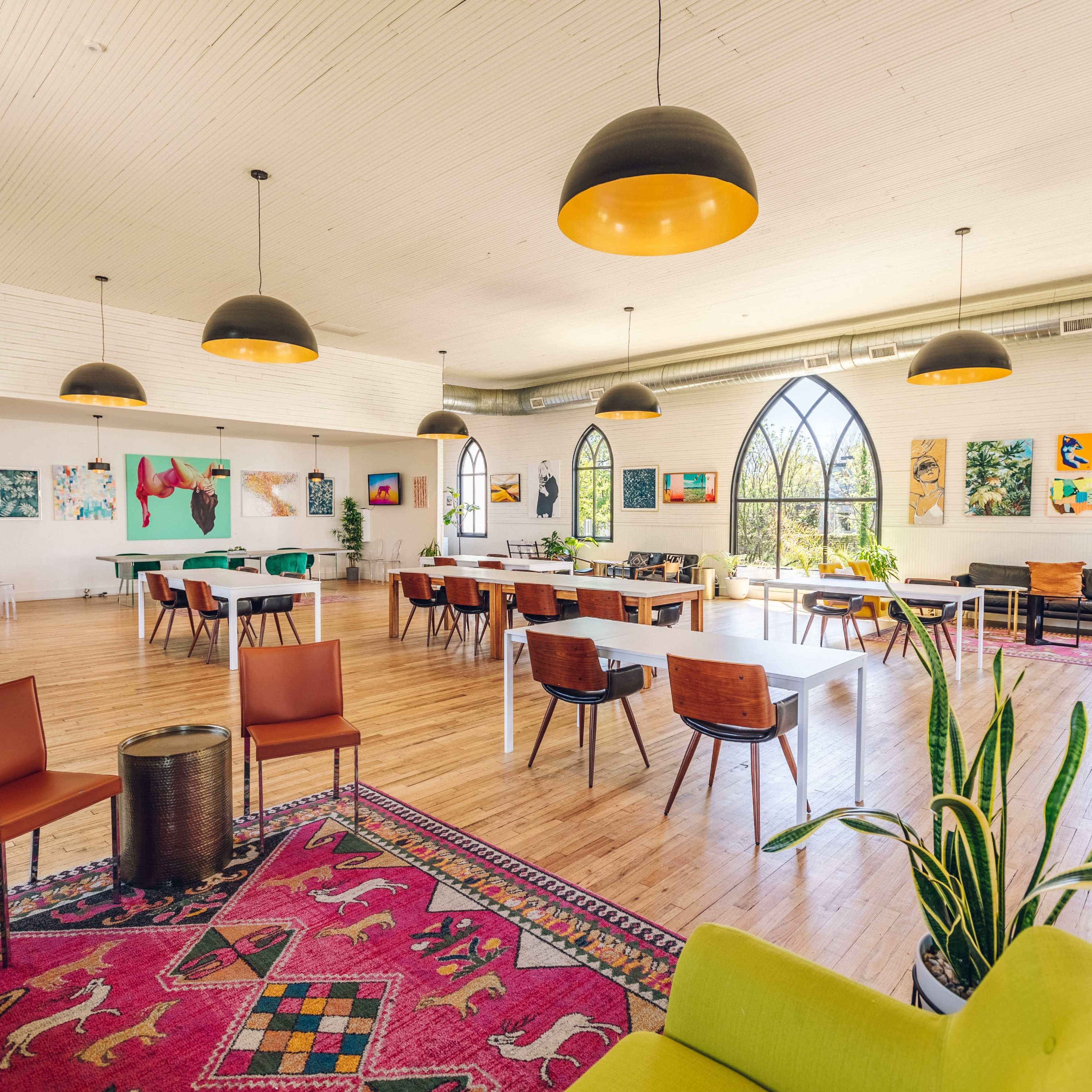 The interior of a spacious, well-lit gathering space features seating areas, artwork on the walls, and large arched windows.