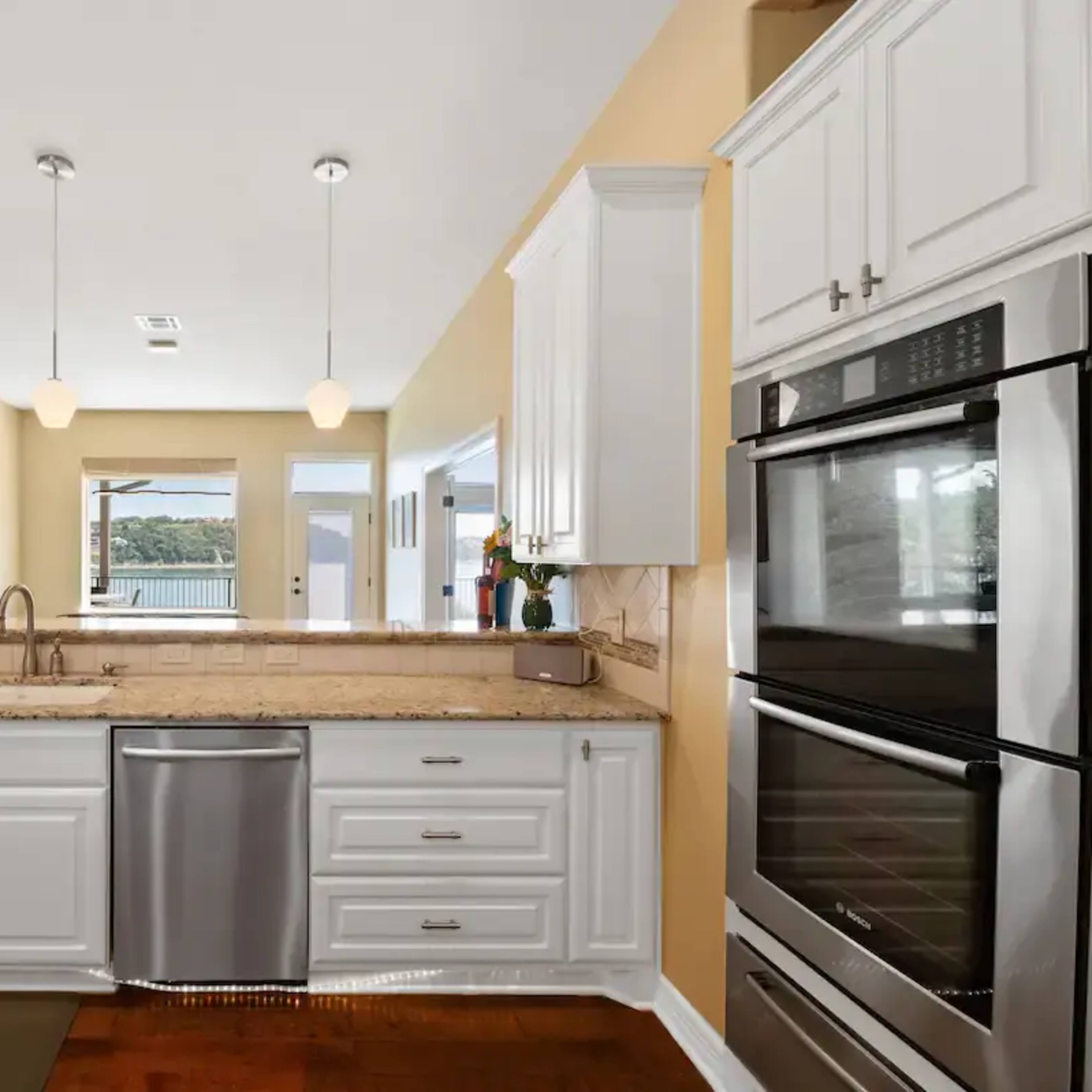 A modern kitchen features white cabinets, stainless steel appliances, and a granite countertop with a view through large windows.